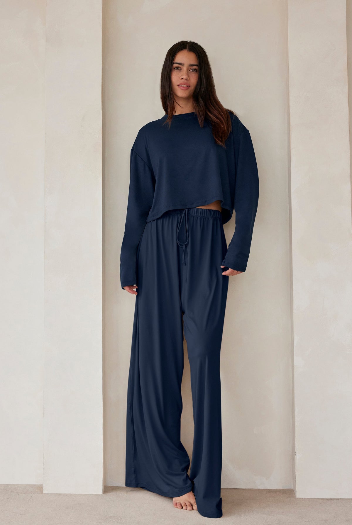 A woman standing against a beige wall in navy lounge pants and a crop long sleeve tee by Bumpsuit