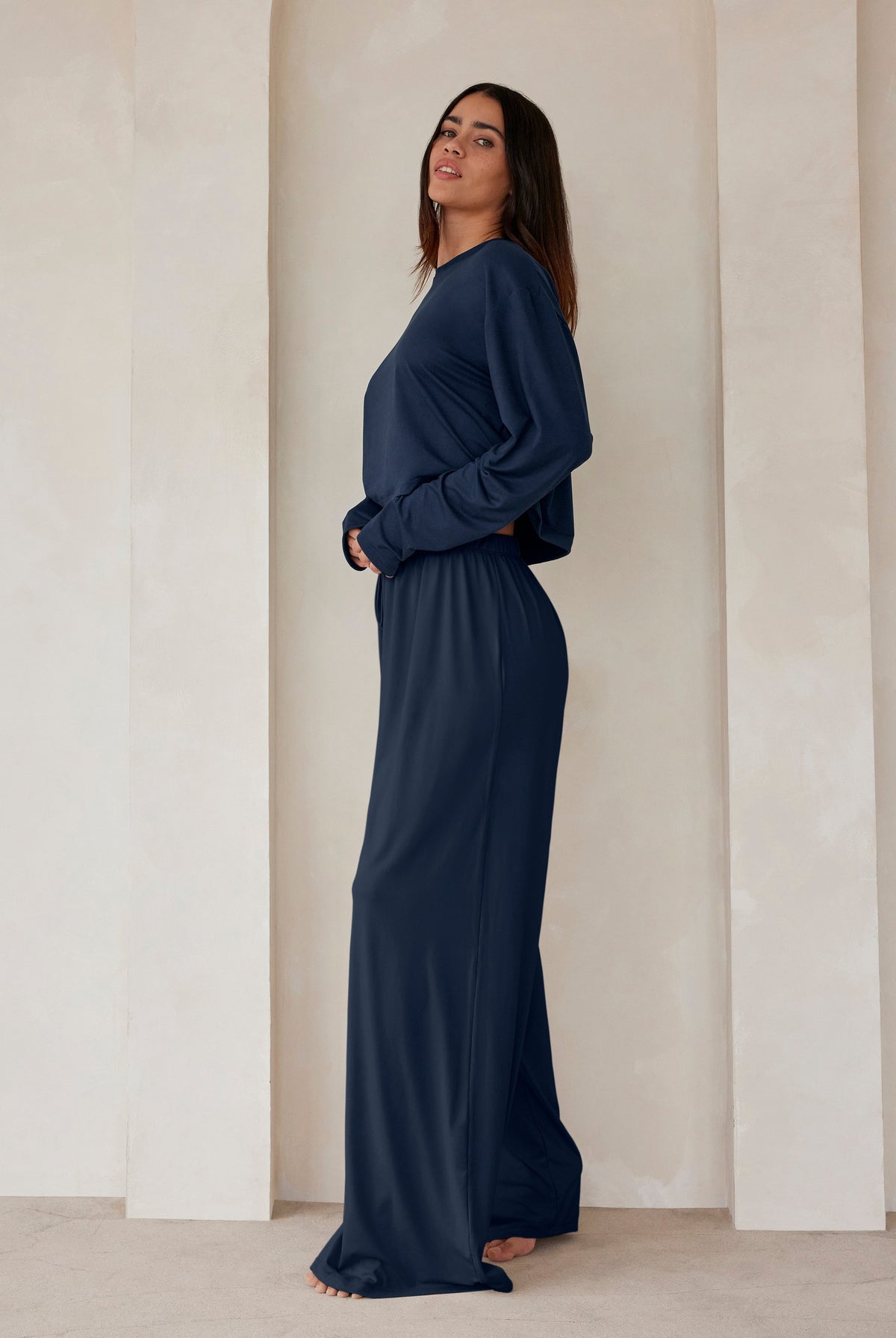 A woman standing against a beige wall in navy lounge pants and a crop long sleeve tee by Bumpsuit