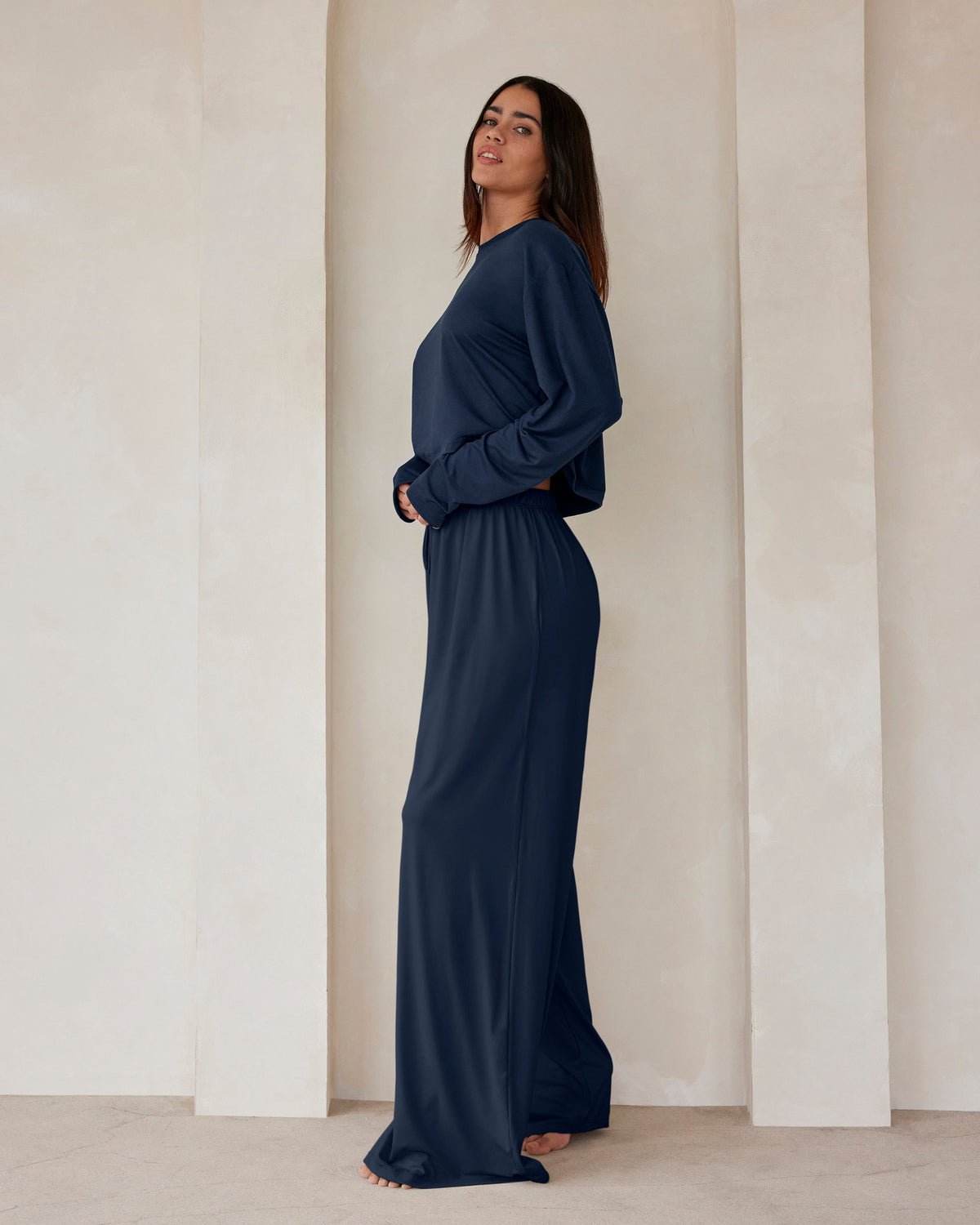 A woman standing against a beige wall in navy lounge pants and a crop long sleeve tee by Bumpsuit