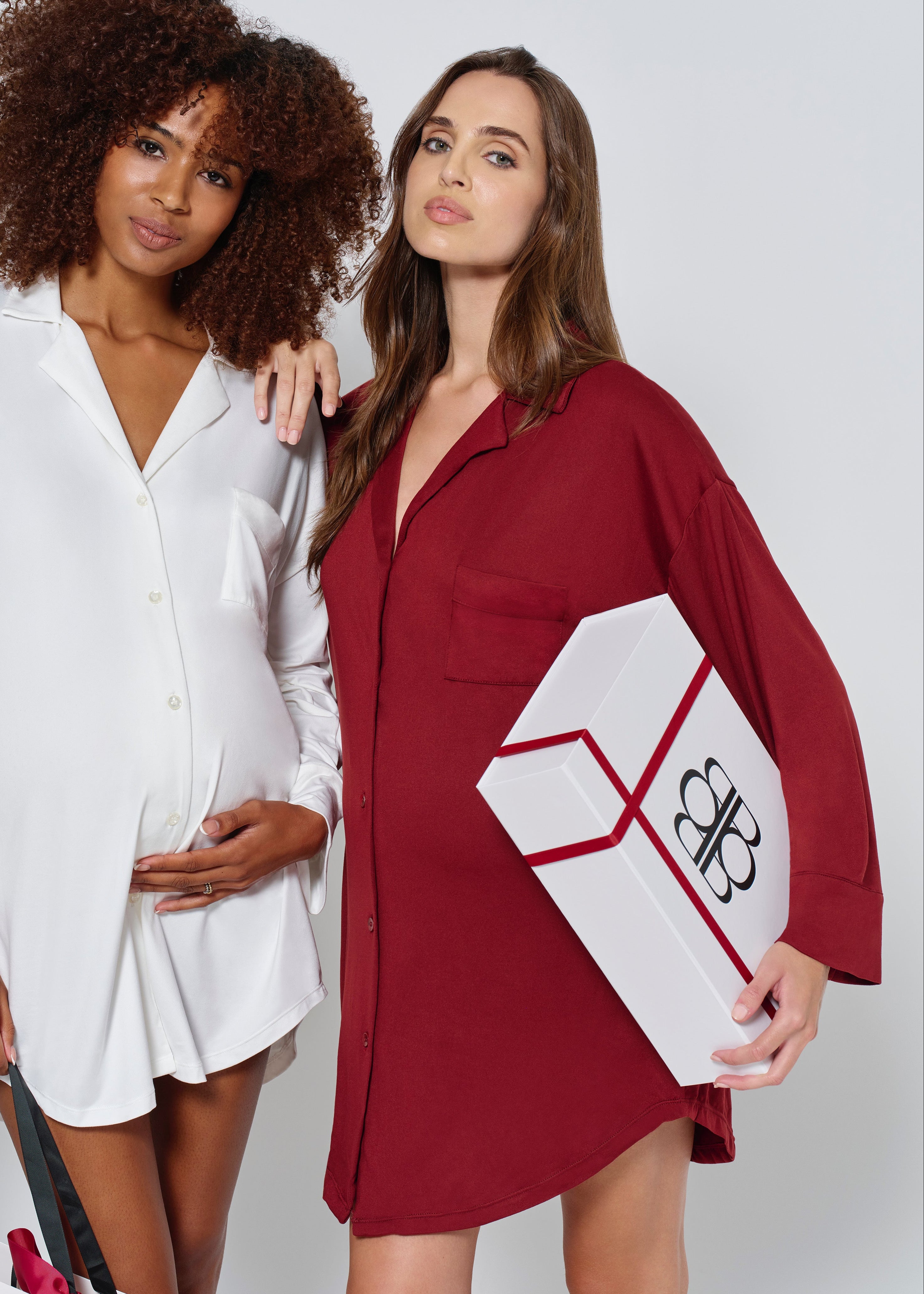Two women posing together, one in a white outfit and the other in a red dress holding a white box with a logo.