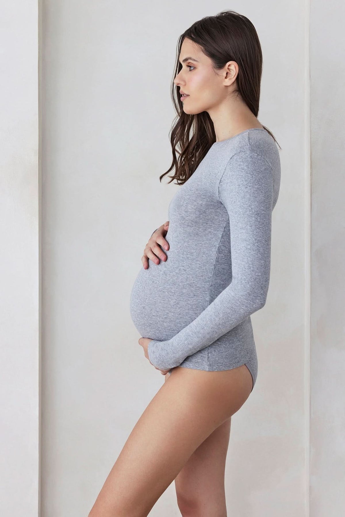 Side view of heather grey maternity cotton rib long sleeve tee showcasing fit over a pregnant belly against a light wall