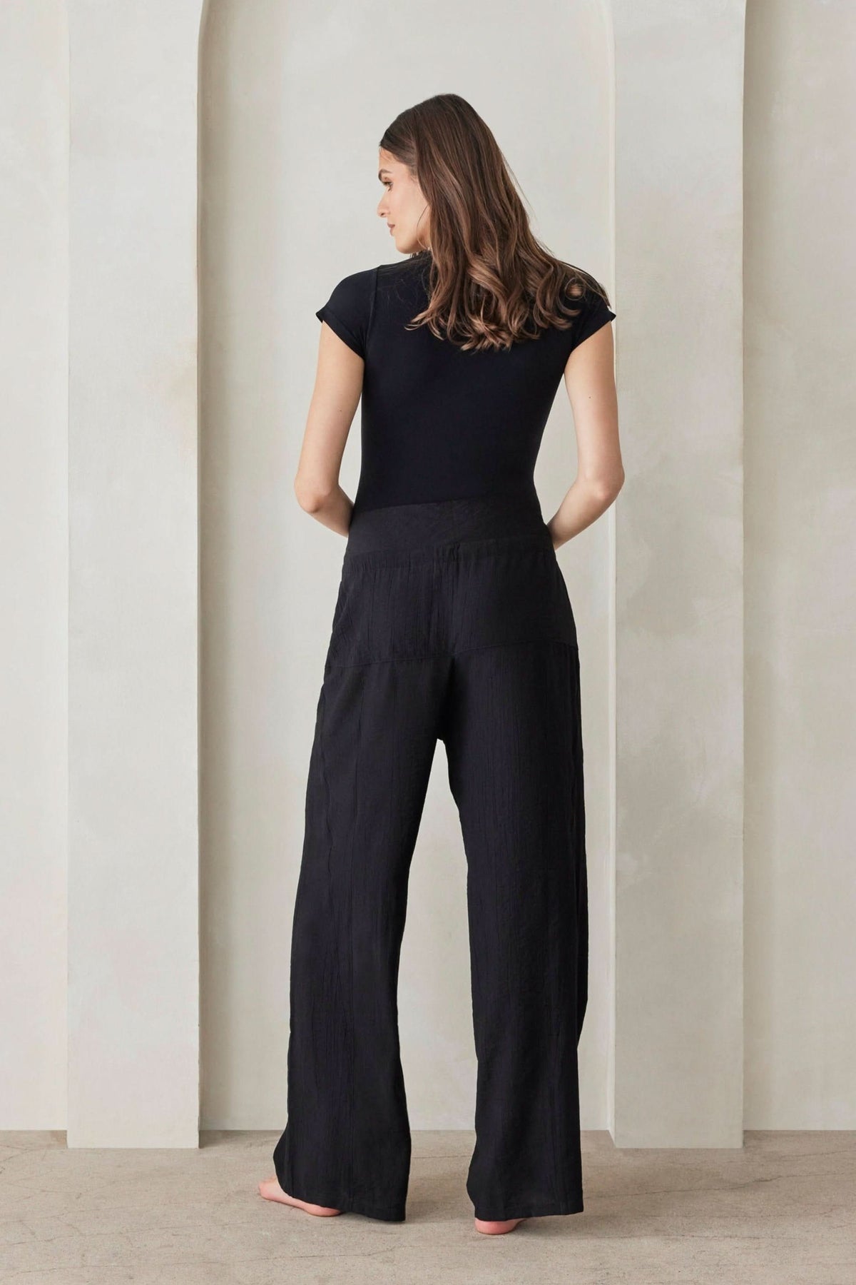 Rear view of The Relaxed Pant in black, showcasing its foldable waistband and wide legs on a model against a light wall