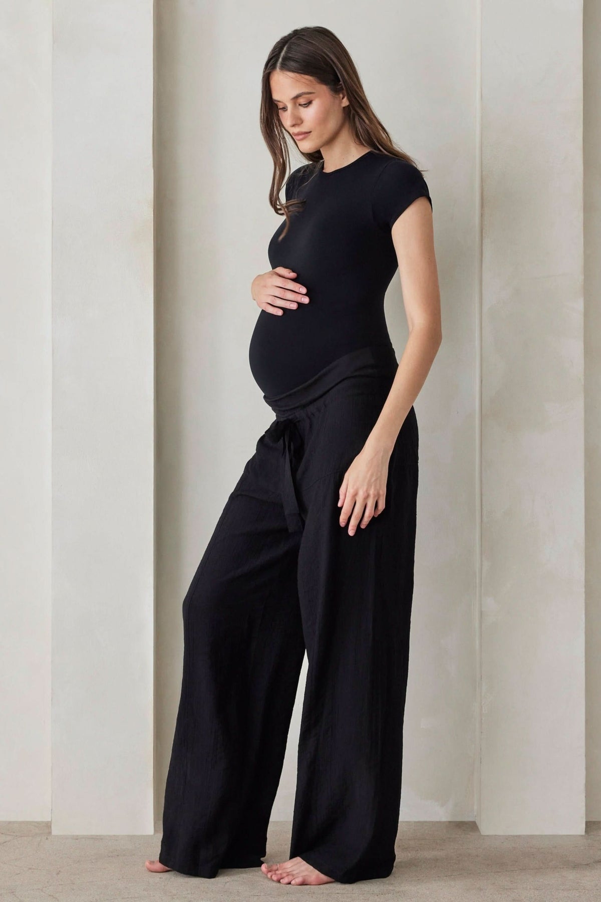 A pregnant woman wearing black relaxed pants and a black shirt by Bumpsuit against a beige feature wall.
