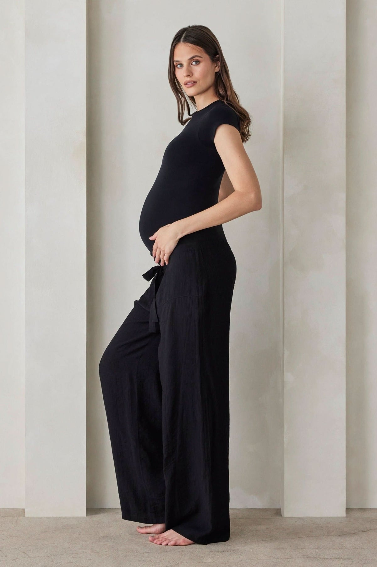 A pregnant woman wearing black relaxed pants and a black shirt by Bumpsuit against a beige feature wall.