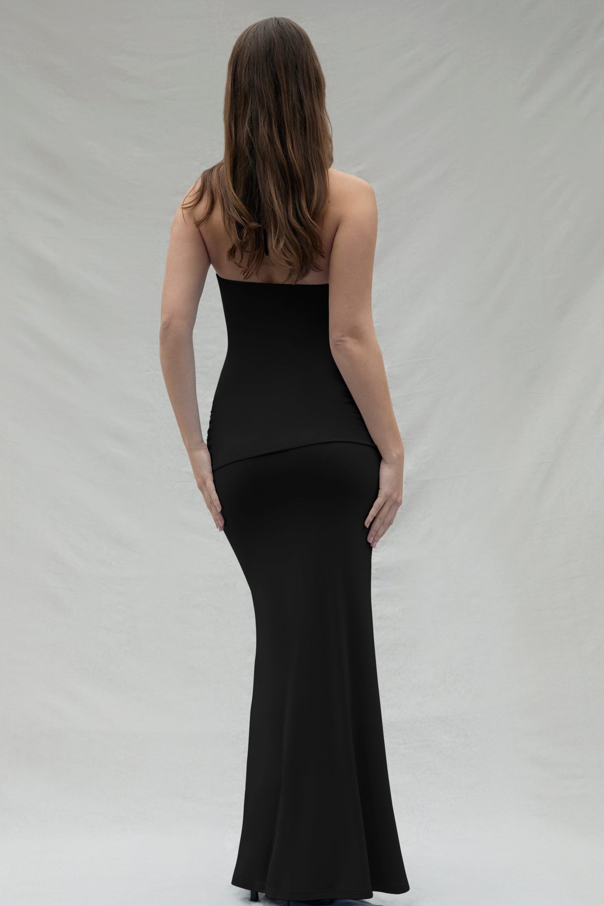 Back view of a woman wearing a black Bumpsuit maternity maxi gown, set against a beige backdrop.