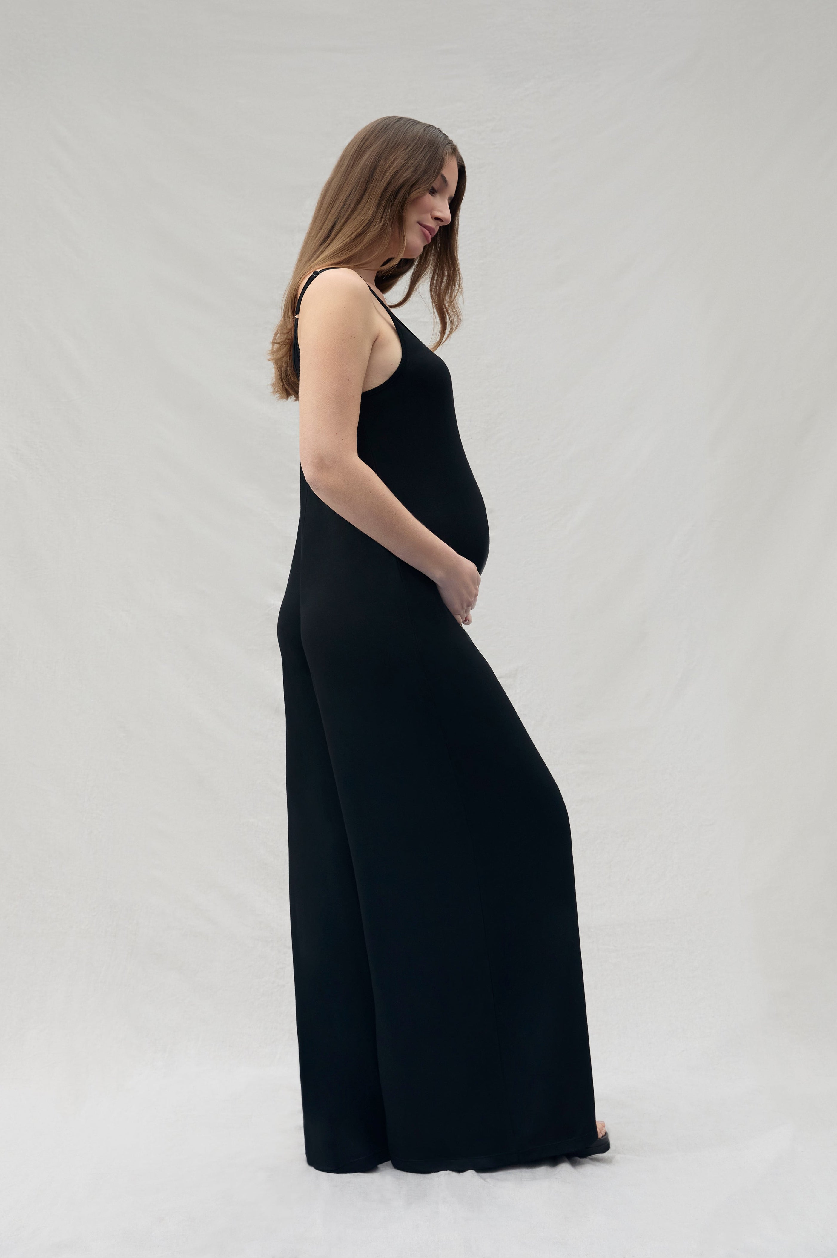 Side view of a women holding under her pregnant belly wearing a cotton black jumpsuit