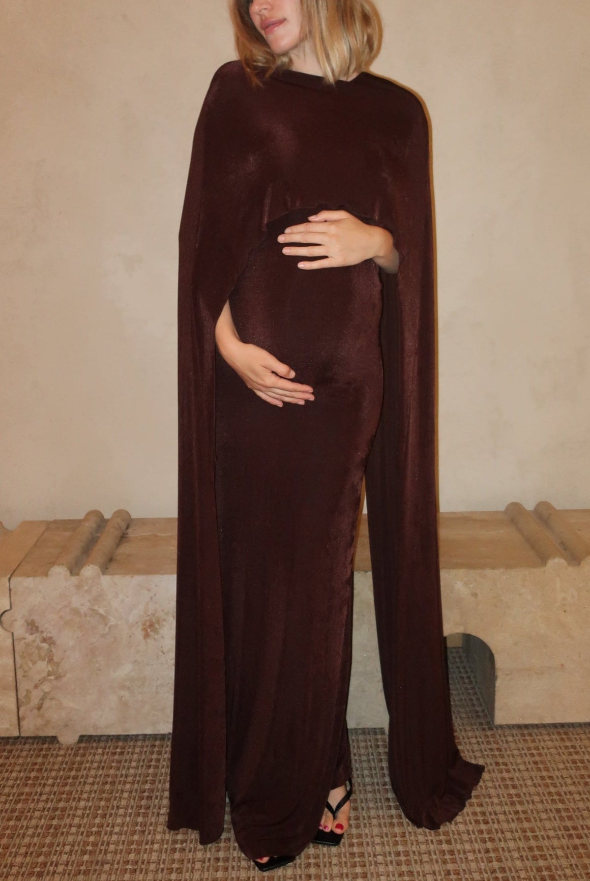 A pregnant woman wearing the Aubrey Bumpsuit maxi dress with a floor length cape detail in chocolate brown against a neutral setting