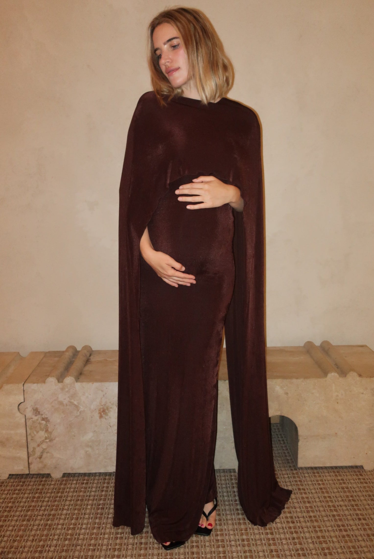 A pregnant woman wearing a Bumpsuit maxi dress with a floor length cape detail in chocolate brown against a neutral setting