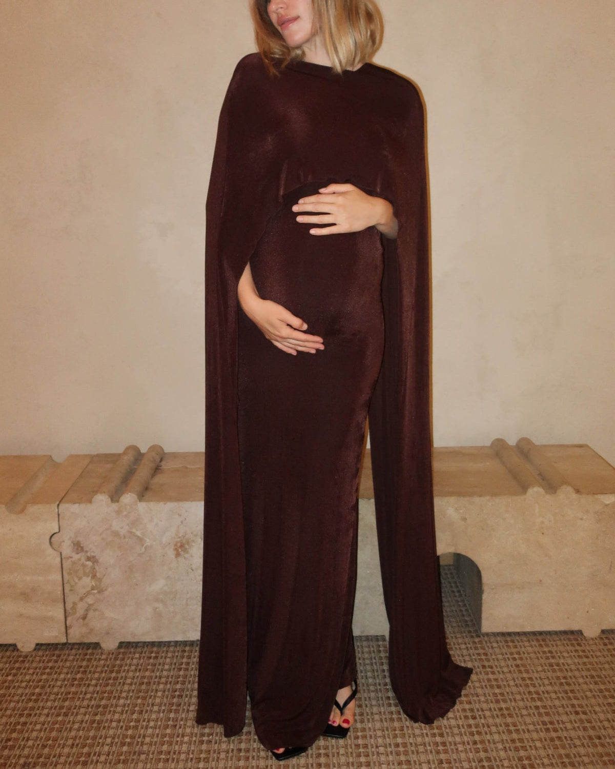 A pregnant woman wearing the Aubrey Bumpsuit maxi dress with a floor length cape detail in chocolate brown against a neutral setting
