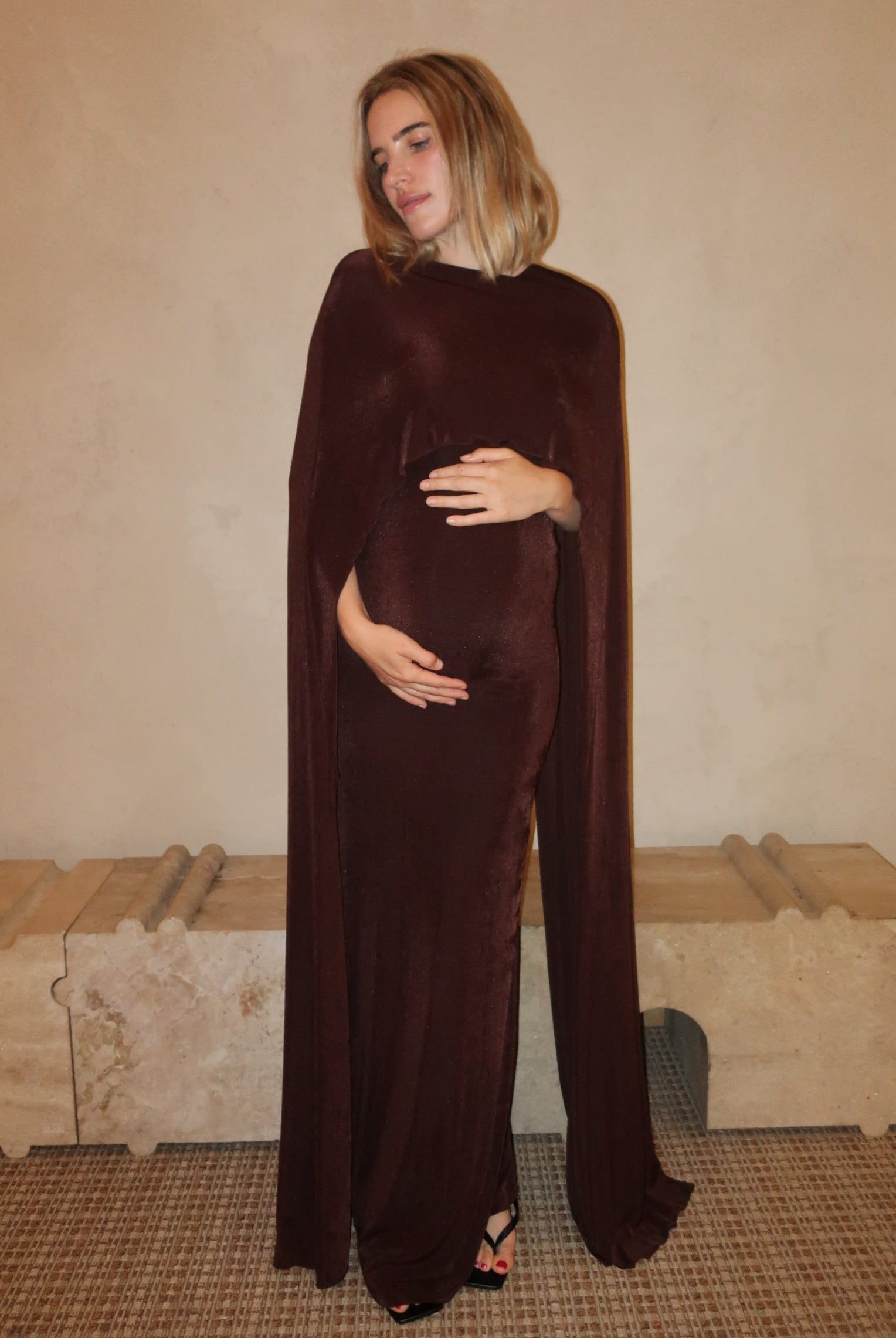 A pregnant woman wearing a Bumpsuit maxi dress with a floor length cape detail in chocolate brown against a neutral setting