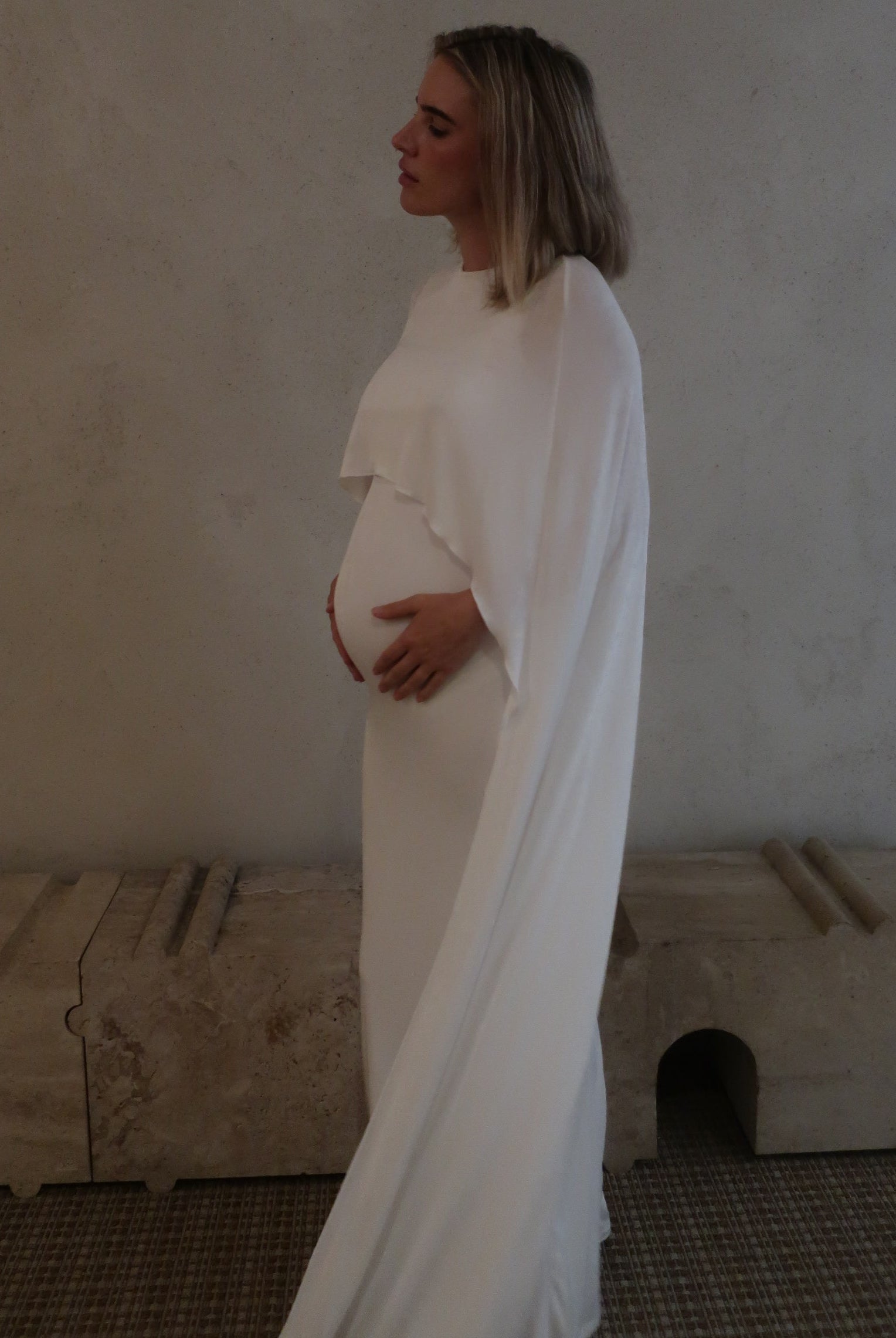 Side view of a pregnant woman wearing a maxi cape style dress in Ivory against a beige background