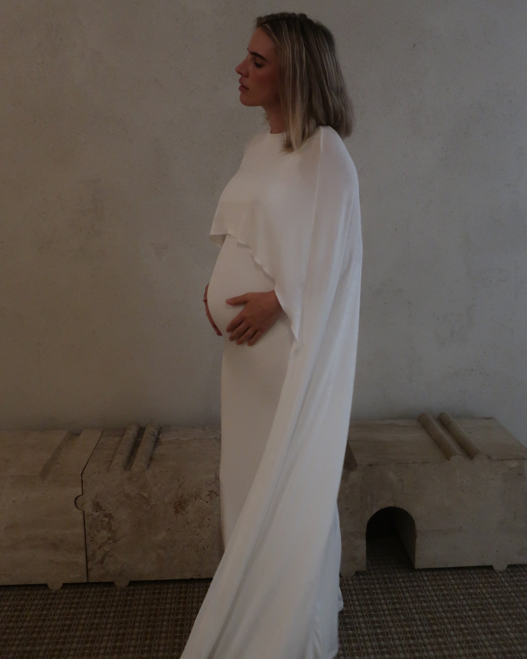 Side view of a pregnant woman wearing a maxi cape style dress in Ivory against a beige background