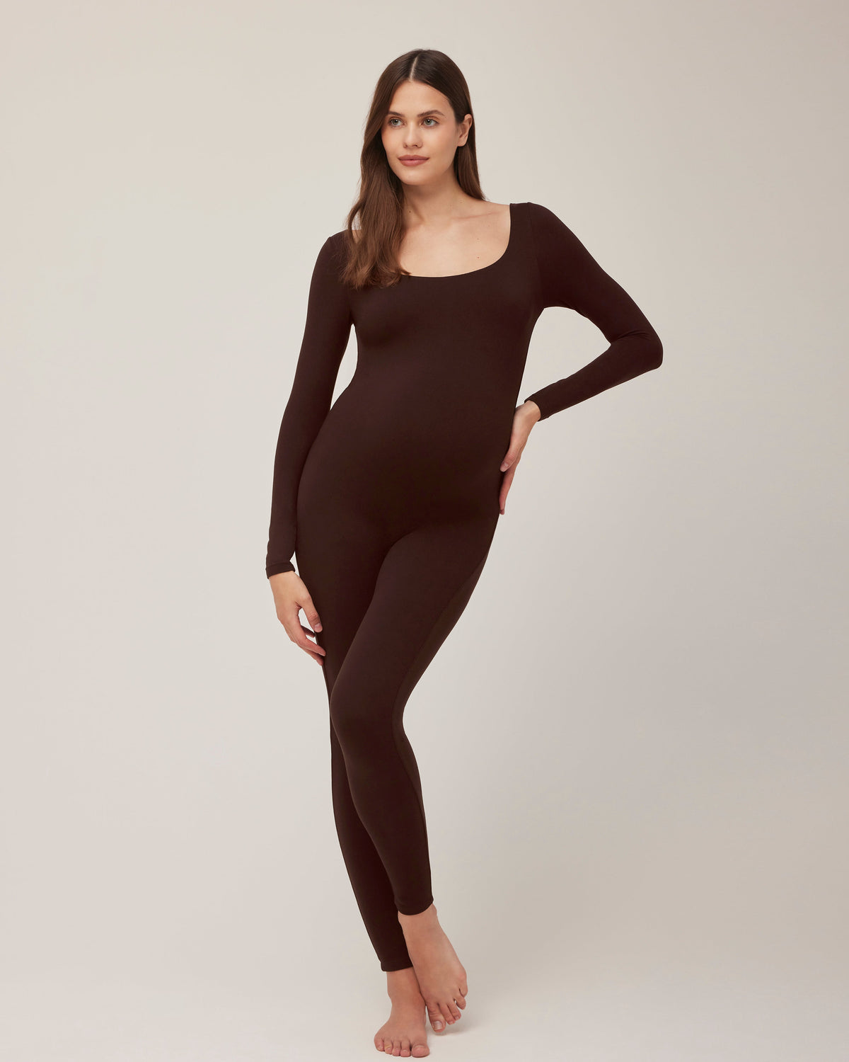 A pregnant woman wearing a Bumpsuit chocolate brown scoop neck long sleeve maternity unitard standing in a studio setting