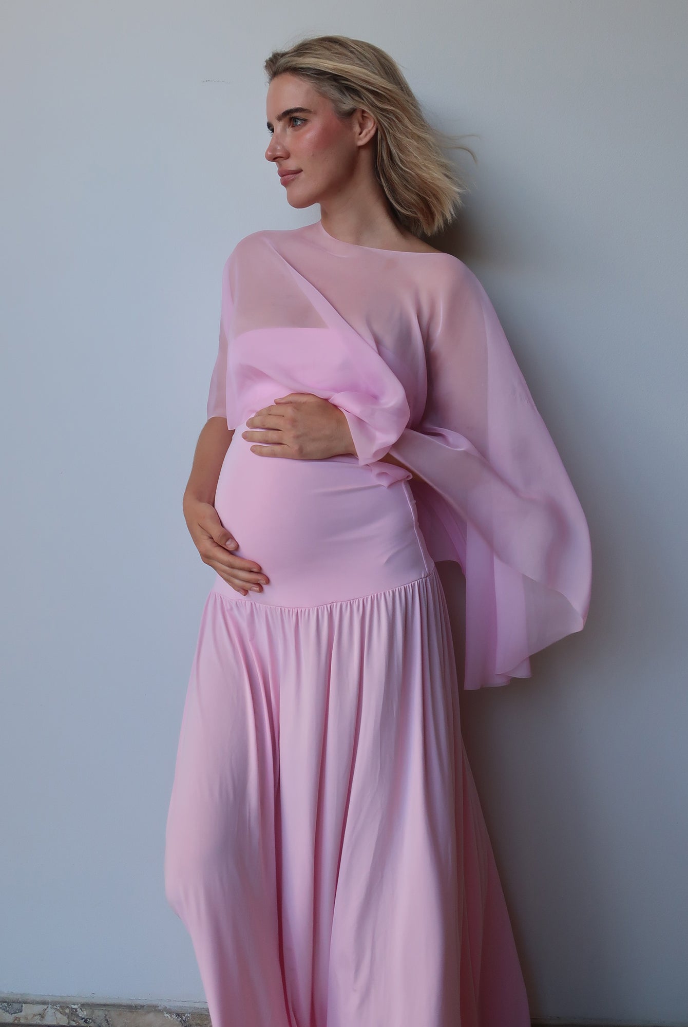 A pregnant woman wearing a light pink maternity dress with a draped chiffon cape, standing against a plain light gray wall.