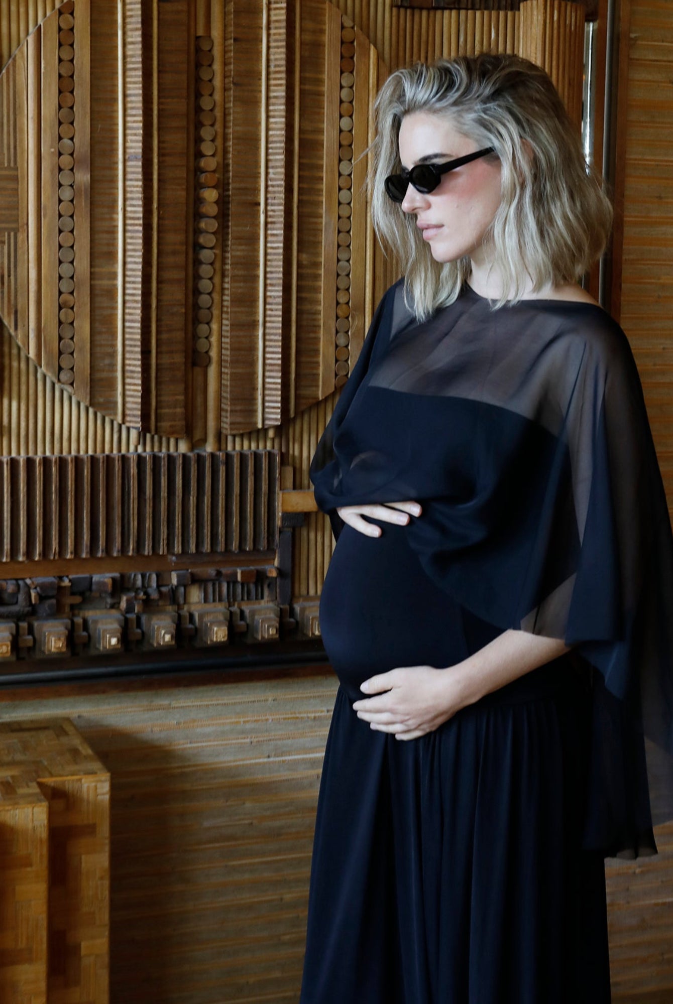 A pregnant woman wearing a black maternity gown with chiffon capelet by Bumpsuit and black sunglasses, standing in front of a wooden geometric wall art piece.
