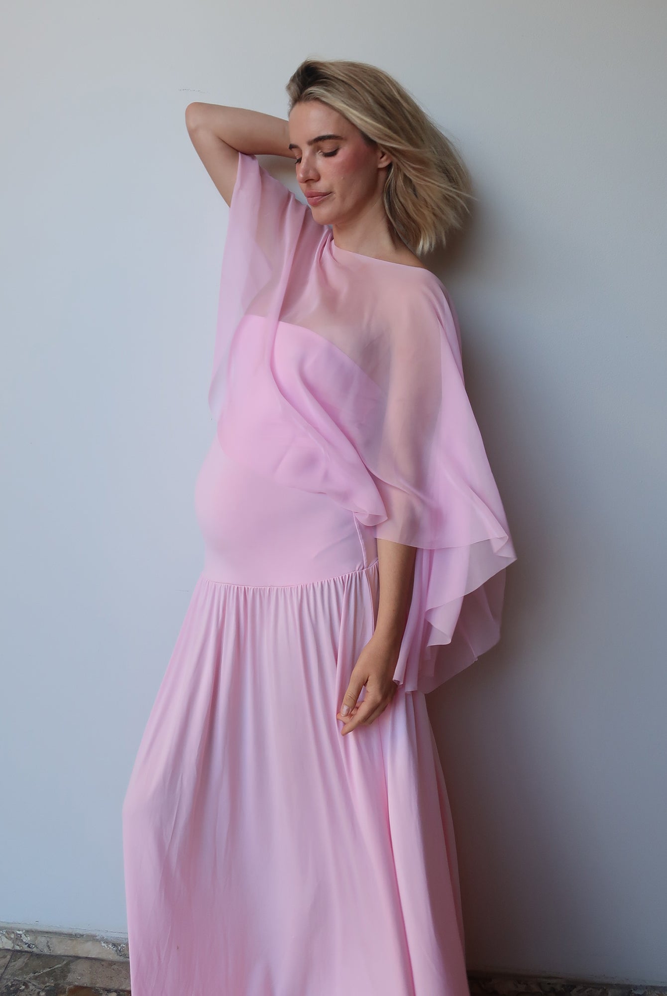 A pregnant woman wearing a Bumpsuit pink maternity dress with a sheer draped cape, posing against a plain light gray wall.