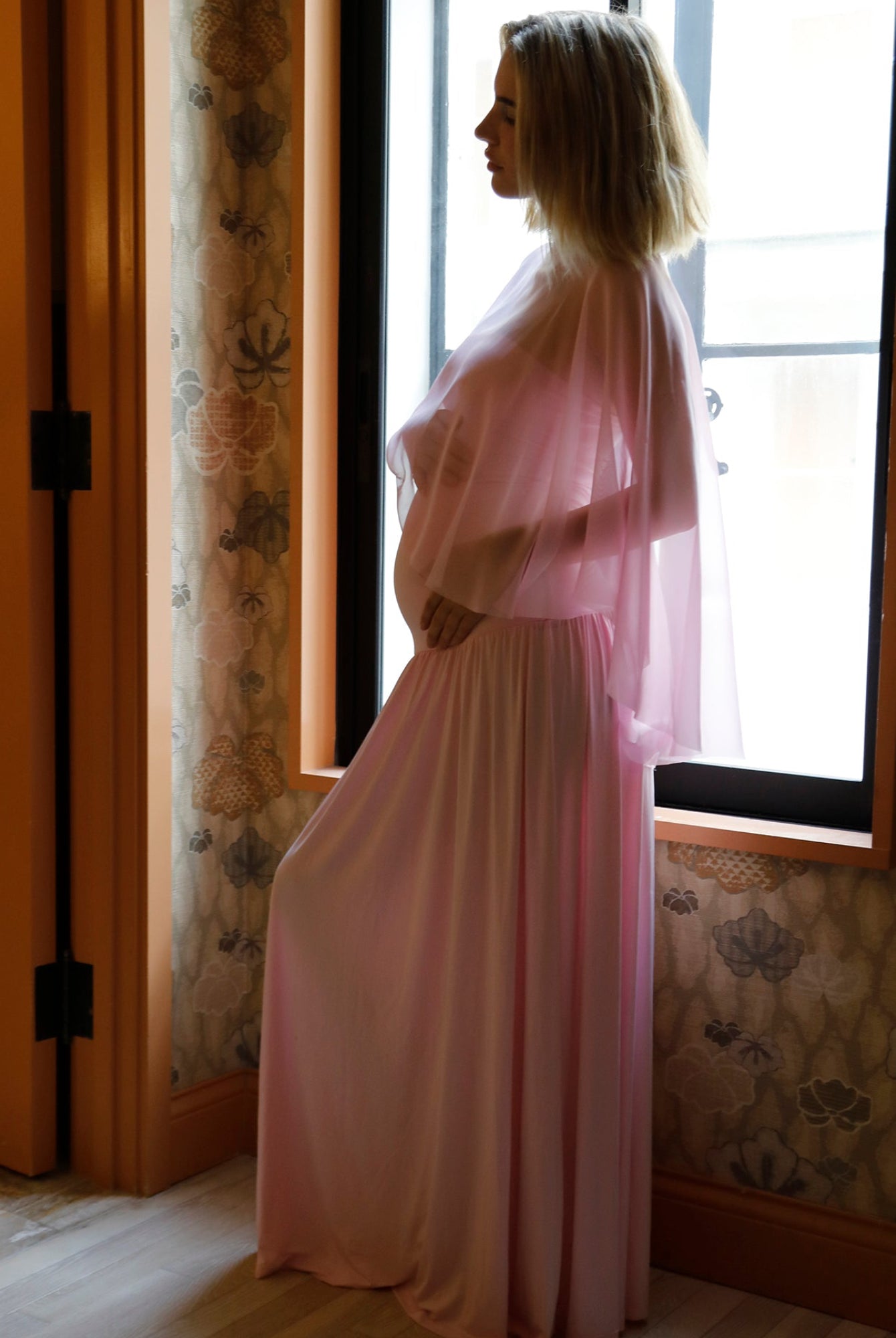 A pregnant woman in a Bumpsuit pink sheer maxi dress with a chiffon cape in profile by a window in a room with floral wallpaper and wood trim.
