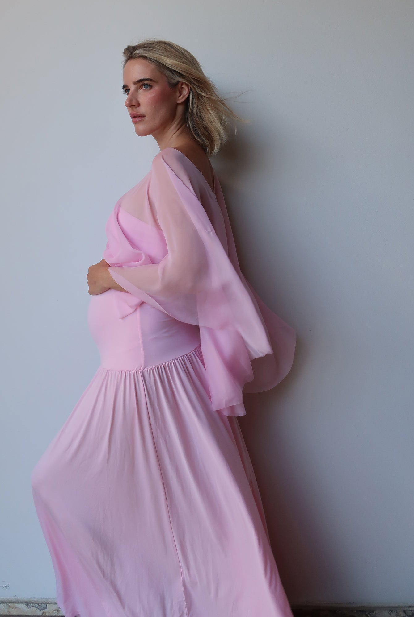 A pregnant woman wearing a flowing light pink maternity dress with chiffon cape by Bumpsuit, standing against a plain light gray wall.