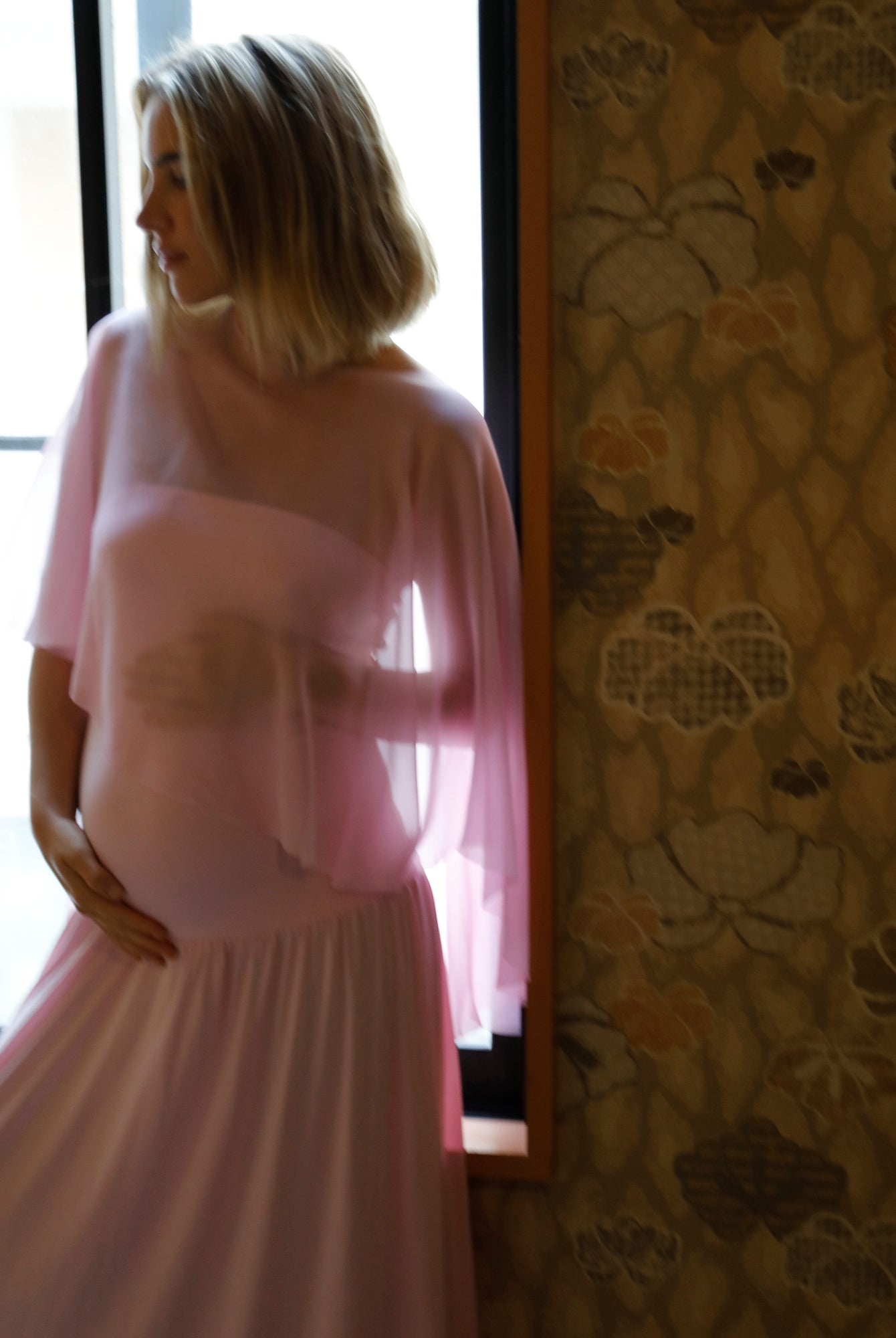 A pregnant woman with blonde hair stands by a window wearing a bumpsuit pink dress with a chiffon cape, infront of a window in a room with patterned wallpaper visible on the right.