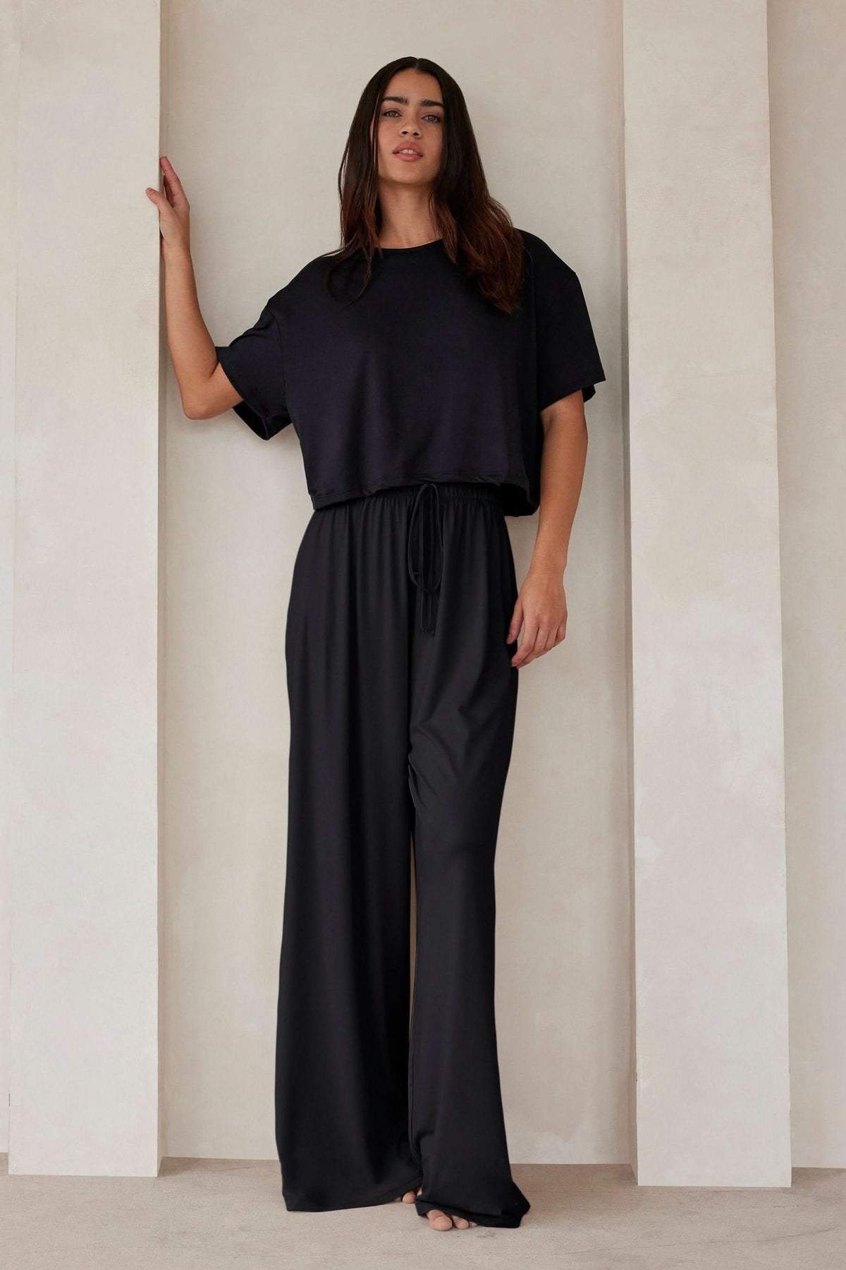 Woman wearing Bumpsuit Maternity The Cloud Short Sleeve Crop Tee in black standing against a beige wall
