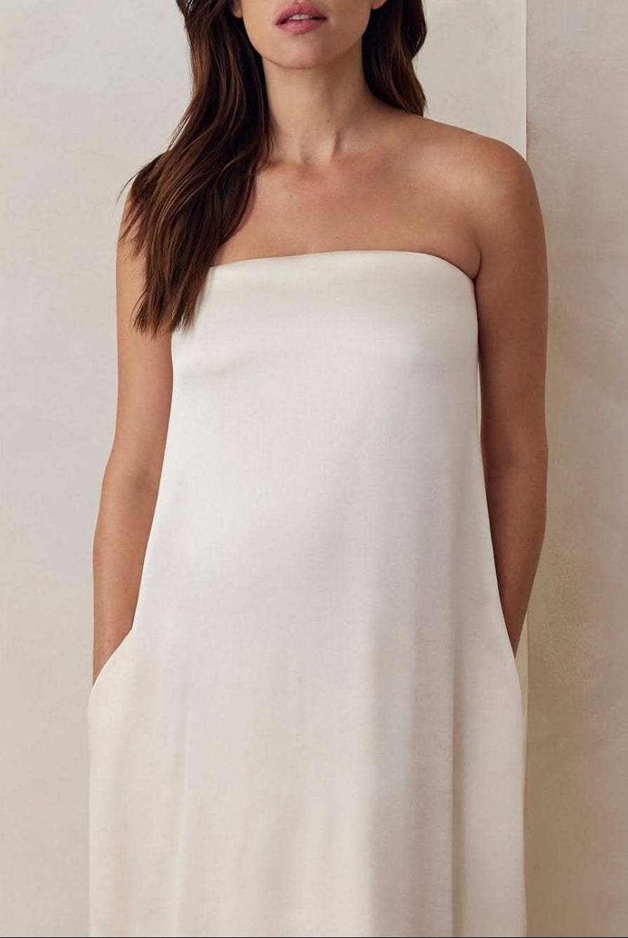 A Pregnant model wearing the Bumpsuit brushed satin gown in ivory against a beige feature wall.