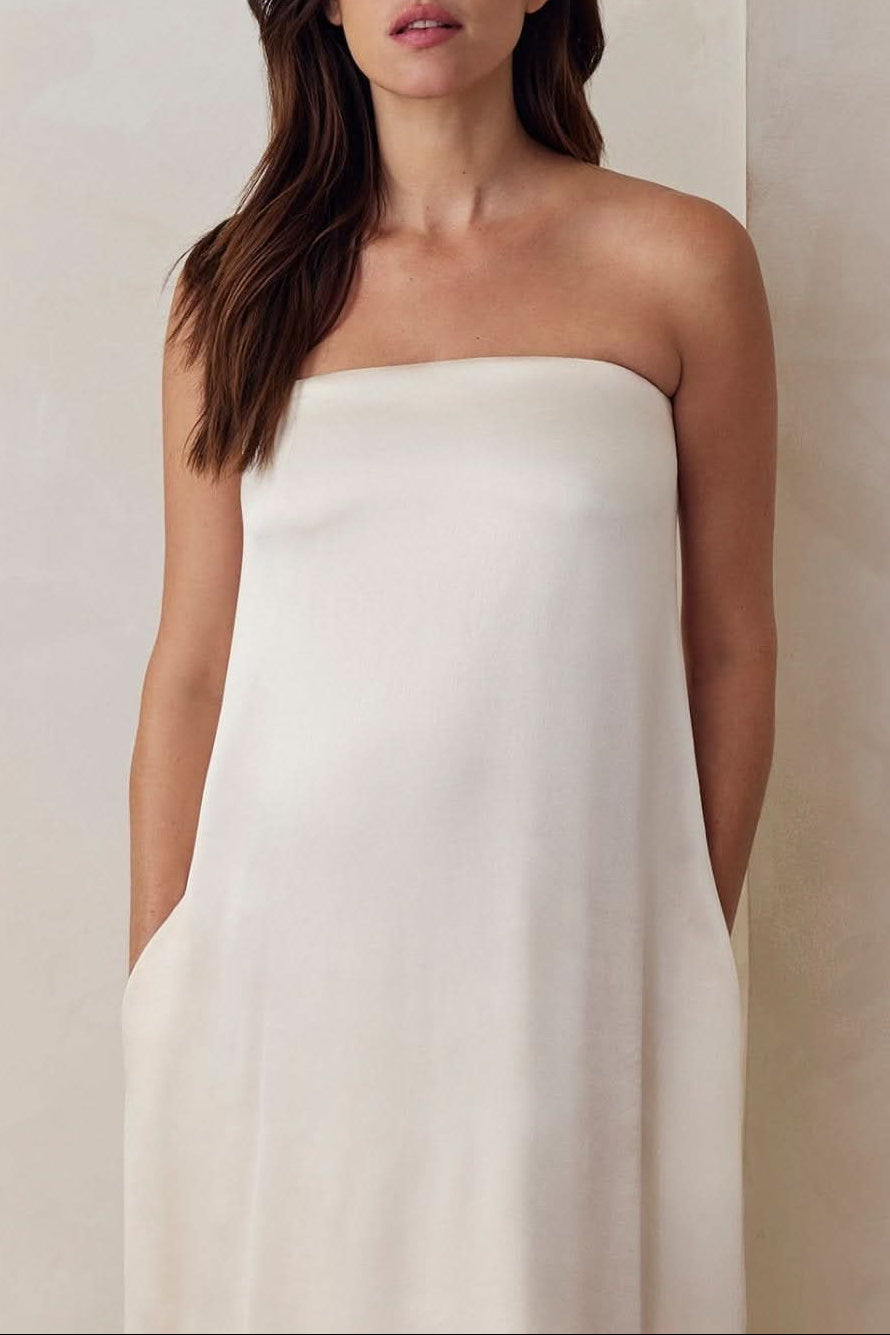 A Pregnant model wearing the Bumpsuit brushed satin gown in ivory against a beige feature wall.