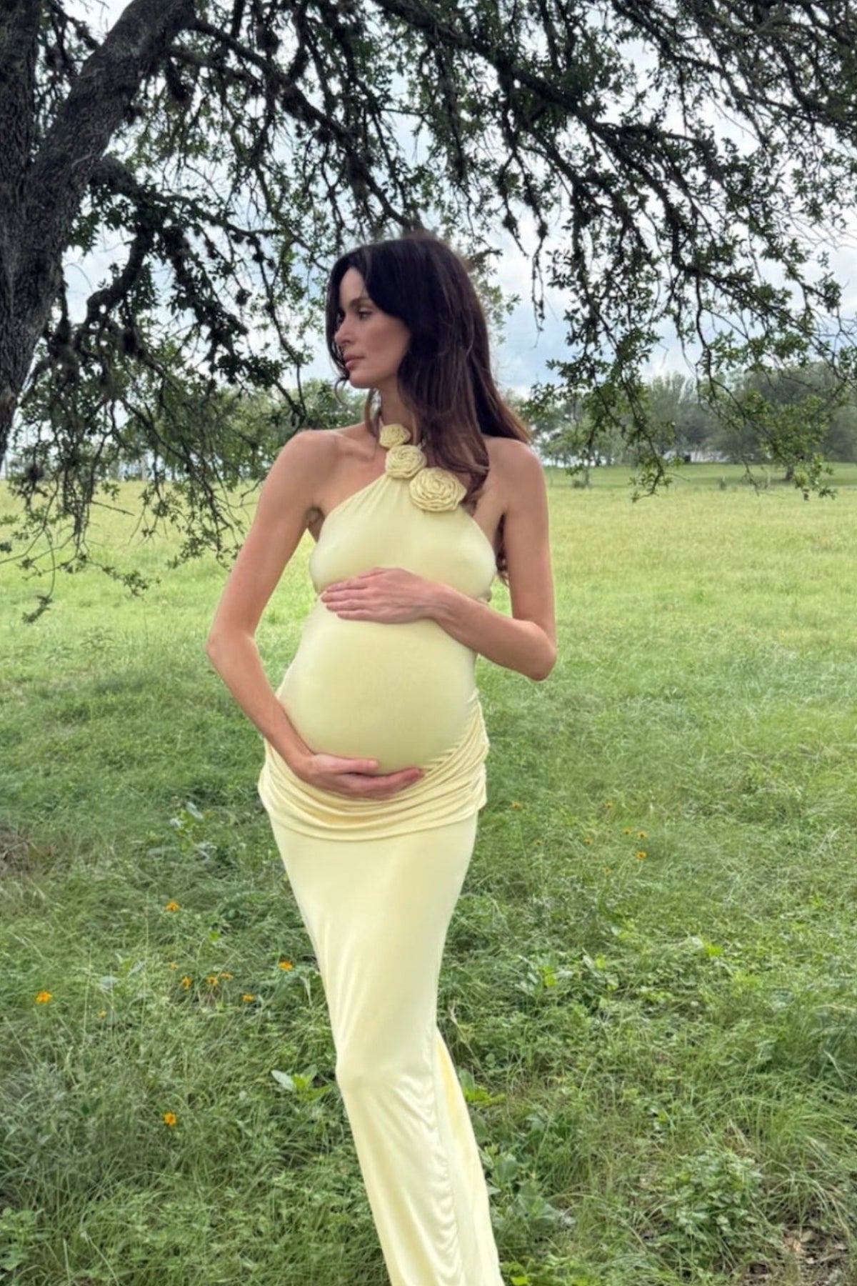 Pregnant woman standing in a grassy field with trees holding her belly wearing a butter yellow bumpsuit maternity dress with a floral halter neckline