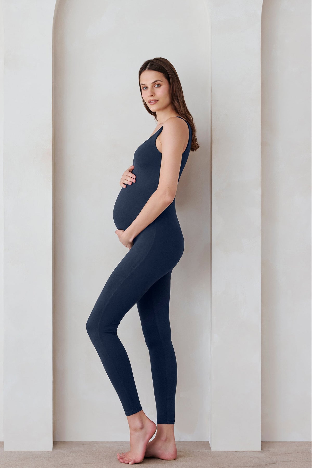 Pregnant woman wearing the Bumpsuit Lucy maternity scoop neck unitard in Navy blue standing against a feature wall