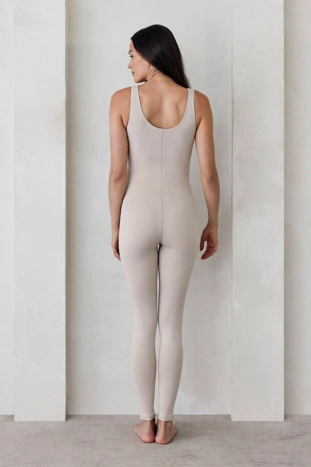 Pregnant woman wearing the Lucy maternity scoop neck unitard in Oat by Bumpsuit set against a feature wall