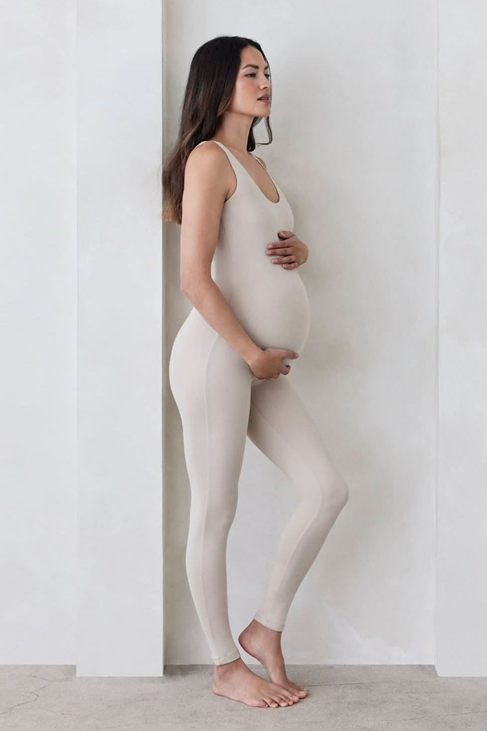 Pregnant woman wearing the Lucy maternity scoop neck unitard in Oat by Bumpsuit set against a feature wall
