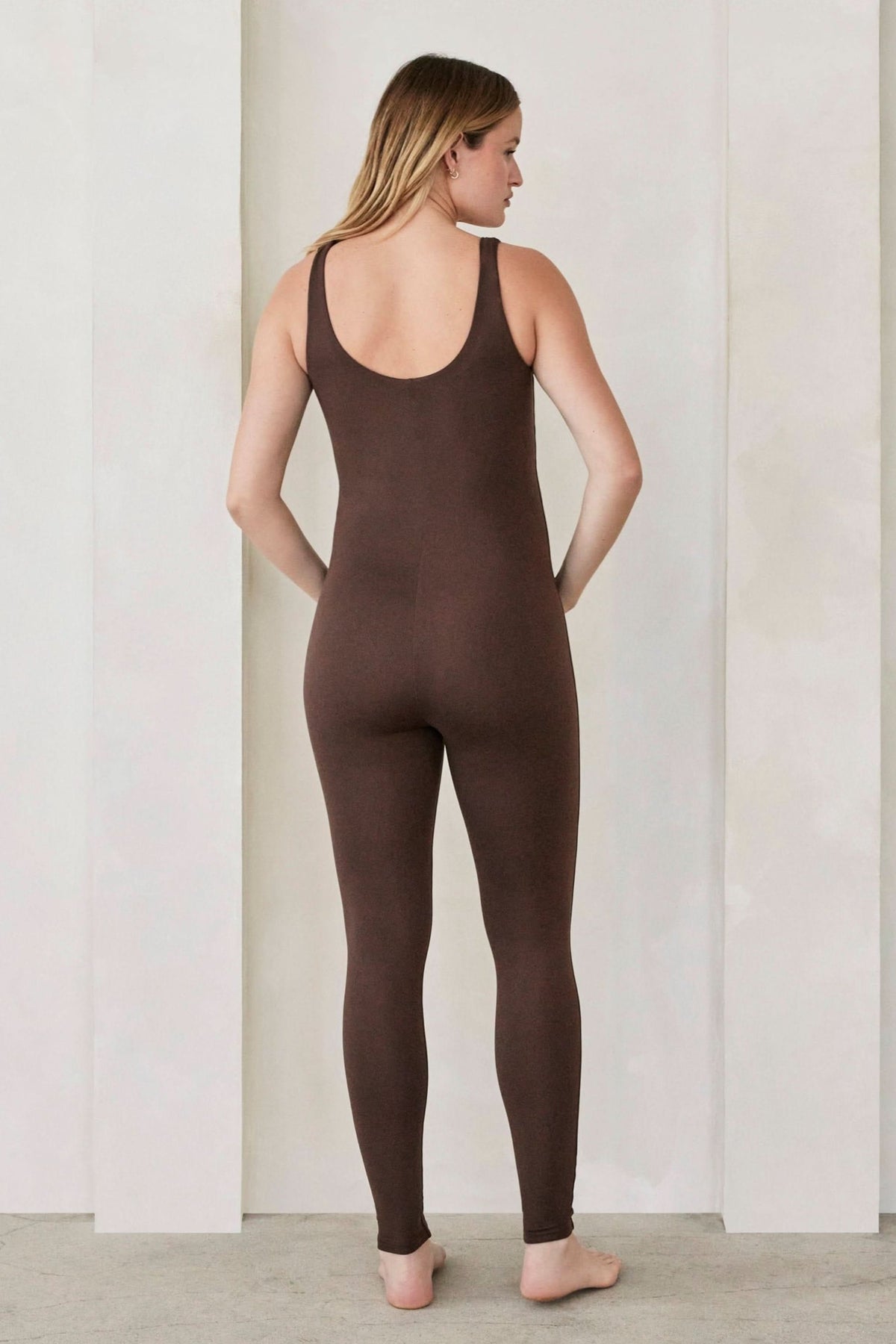 Pregnant woman wearing the lucy maternity unitard in chocolate brown by bumpsuit set against a feature wall