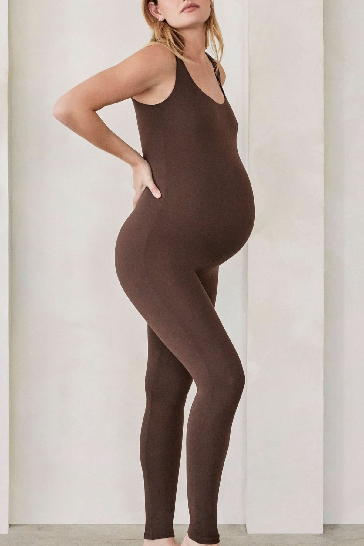 Pregnant woman wearing the lucy maternity unitard in chocolate brown by bumpsuit set against a feature wall