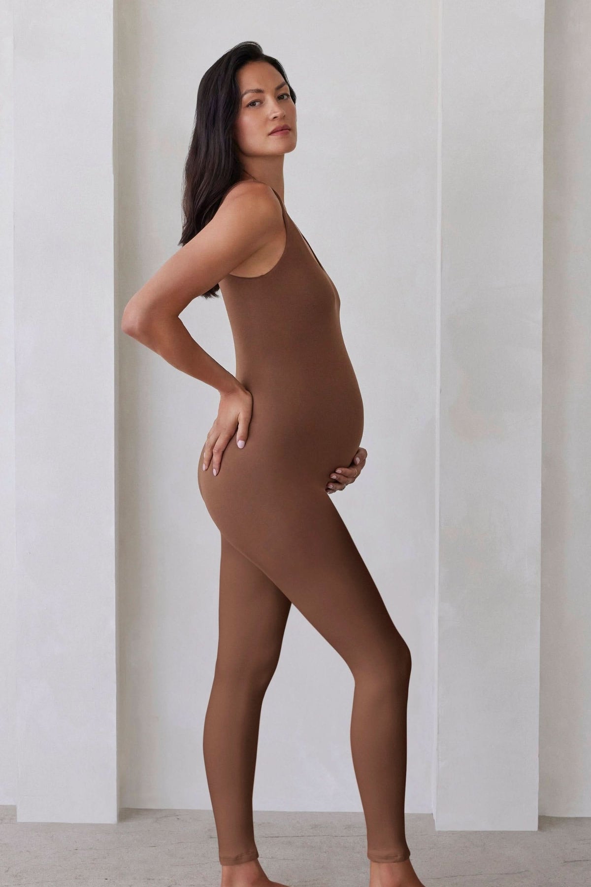 Pregnant woman wearing the Lucy maternity scoop neck unitard in Mocha by Bumpsuit set against a feature wall