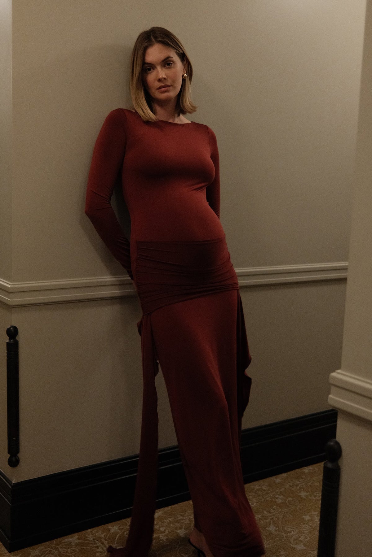 A pregnant woman wearing a long-sleeve, boat neck, floor-length burgundy Bumpsuit maternity dress with a gathered waist, posing in a hallway with beige walls and dark trim.