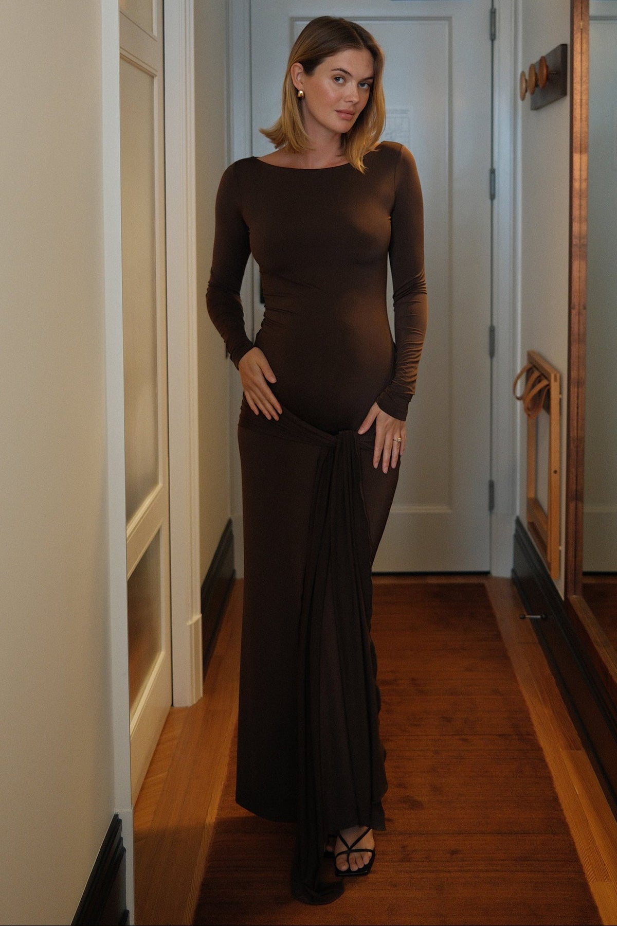A woman wearing a long-sleeve, chocolate brown maternity maxi dress with a gathered, draped waist tie detail, standing in a hallway with wooden floors.
