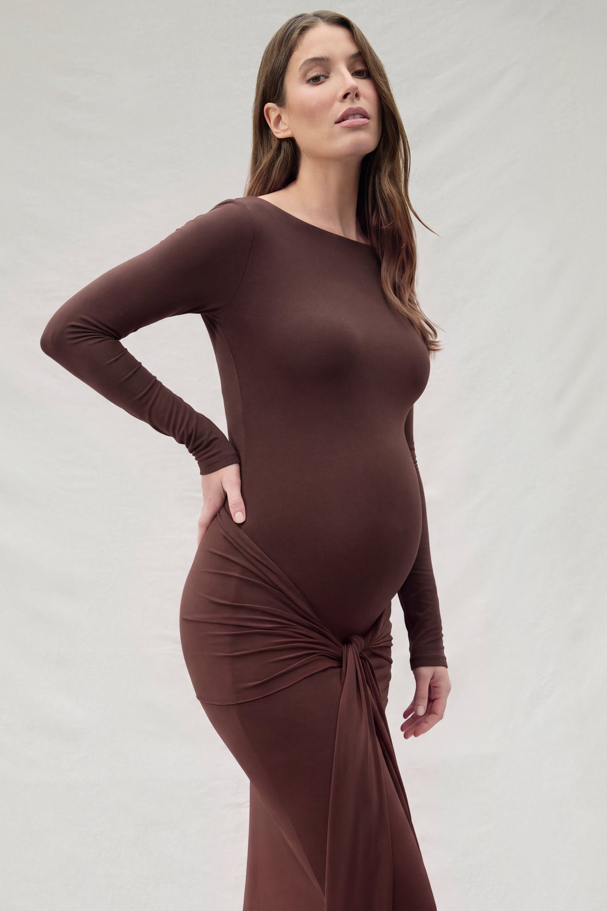 Side view of a pregnant woman wearing a Bumpsuit maternity chocolate brown, maxi dress with a tie feature at the waist.