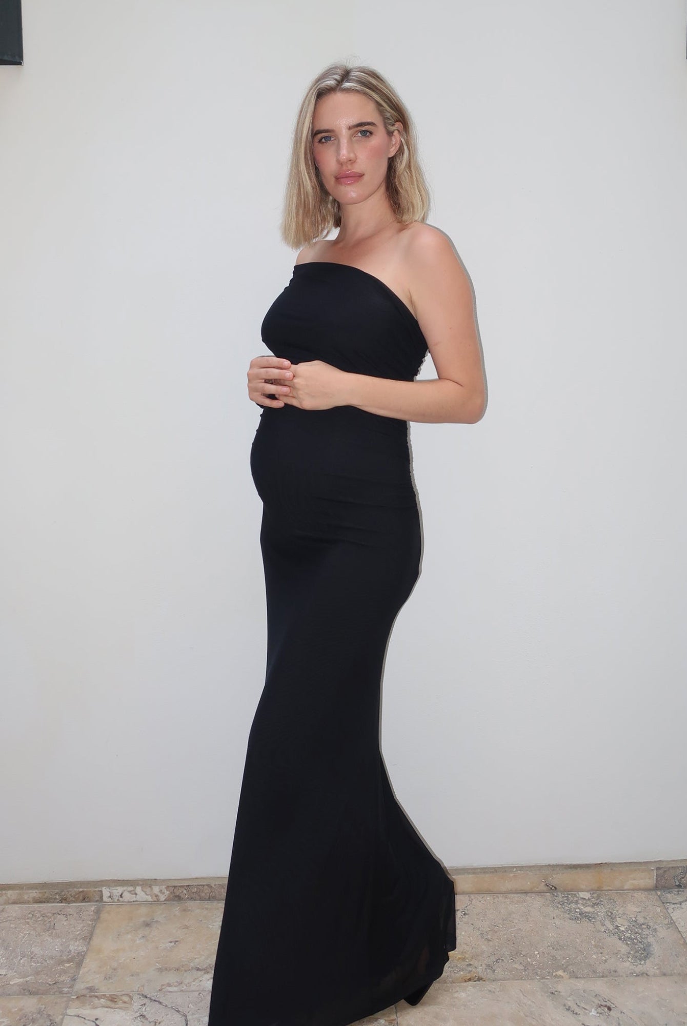 A pregnant woman wearing the Vanessa Bumpsuit off the shoulder gown, standing against a white wall with stone tile flooring.