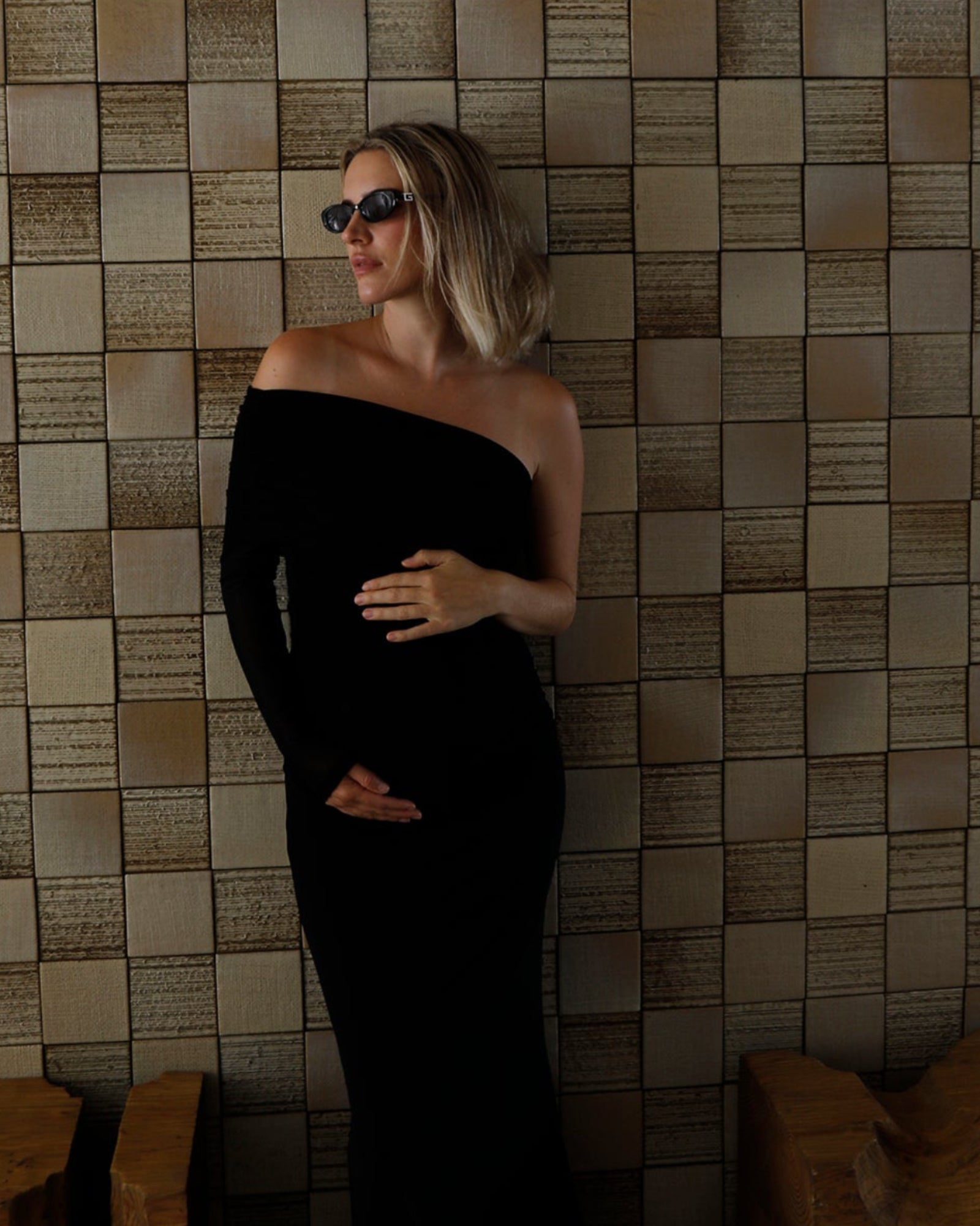 A pregnant woman wearing a Bumpsuit black off-the-shoulder floor-length gown and dark sunglasses stands against a beige tiled wall with wooden benches at the bottom.