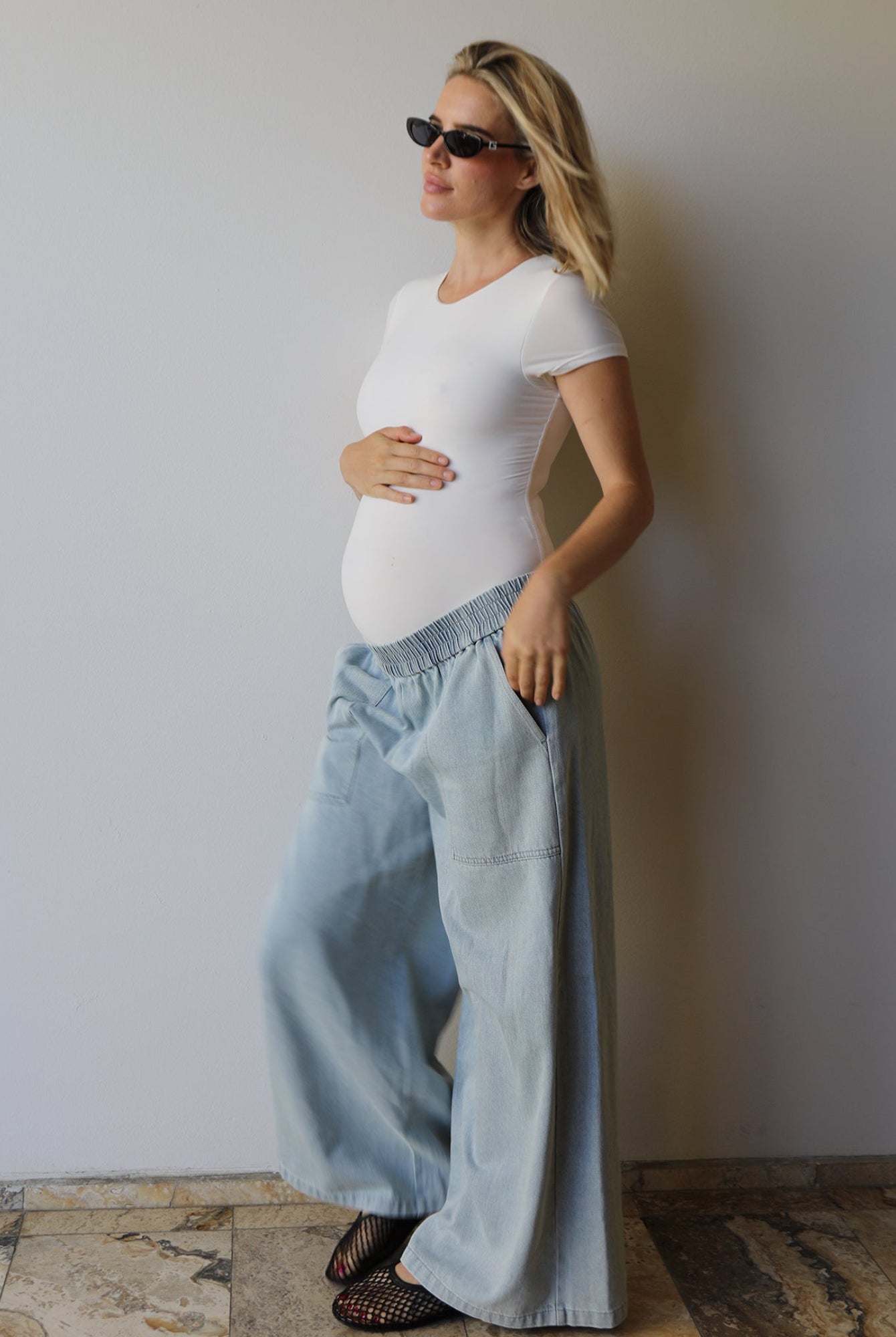A pregnant woman wearing a Bumpsuit white short-sleeve everyday shirt and light blue wide-leg denim pants, standing against a plain light gray wall on a stone-tiled floor.