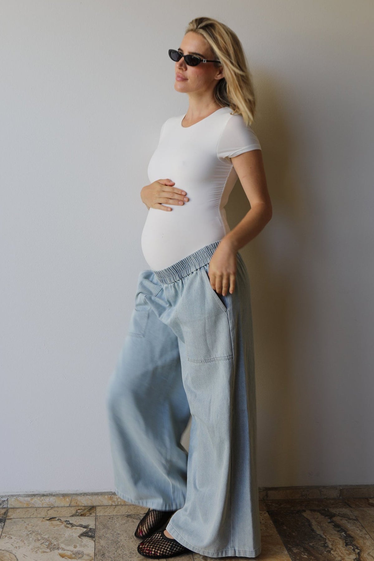 A pregnant woman wearing a Bumpsuit white short-sleeve everyday shirt and light blue wide-leg denim pants, standing against a plain light gray wall on a stone-tiled floor.