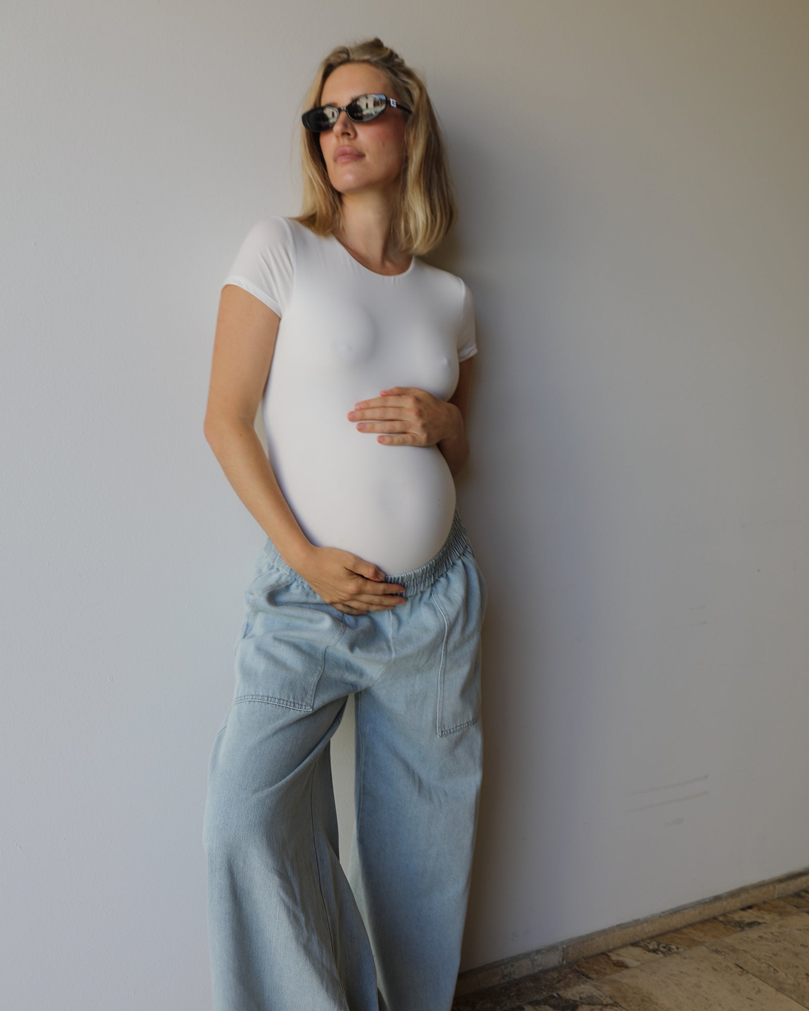 A pregnant woman wearing a fitted white short-sleeve t-shirt and light blue wide-leg jeans, standing against a plain light gray wall.