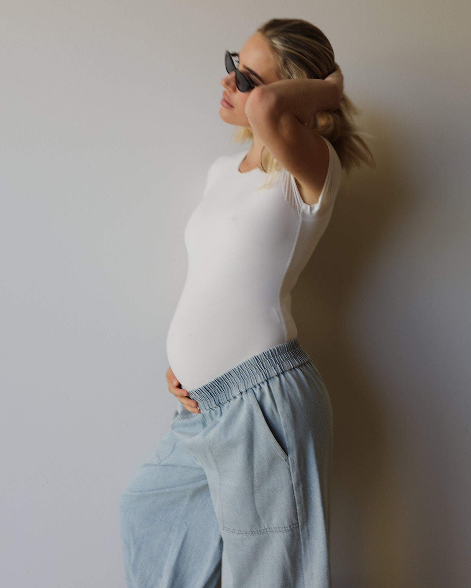 A pregnant woman wearing a white short-sleeve bodysuit and light blue wide-leg jeans by Bumpsuit, posing in profile against a plain light gray wall.