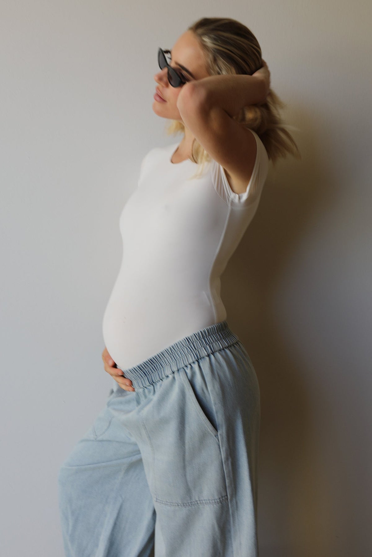A pregnant woman wearing a white short-sleeve bodysuit and light blue wide-leg jeans by Bumpsuit, posing in profile against a plain light gray wall.