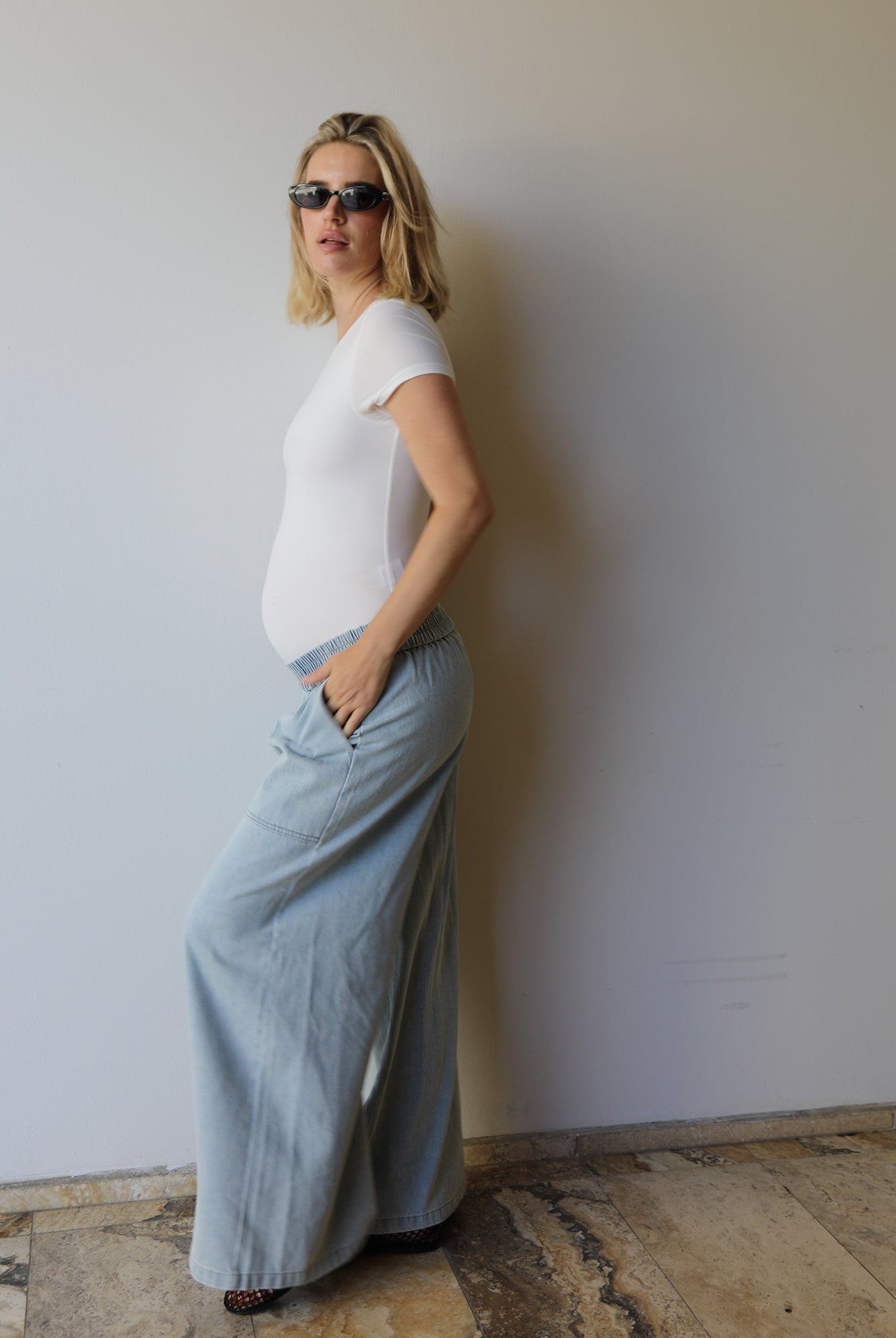 A pregnant woman wearing a white short-sleeve t-shirt and light blue wide-leg denim maxi jeans by Bumpsuits, standing against a plain off-white wall.