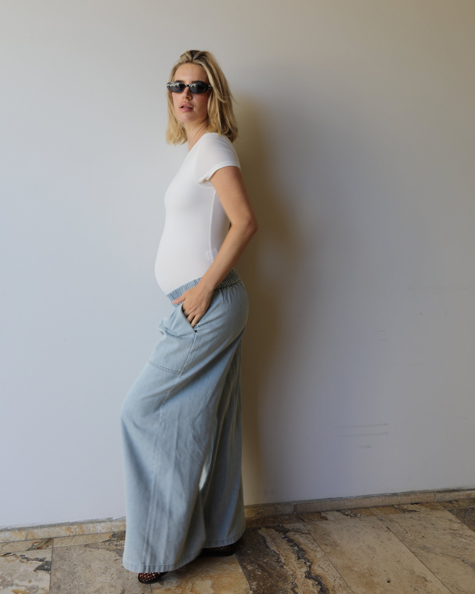 A pregnant woman wearing a white short-sleeve t-shirt and light blue wide-leg denim maxi jeans by Bumpsuits, standing against a plain off-white wall.