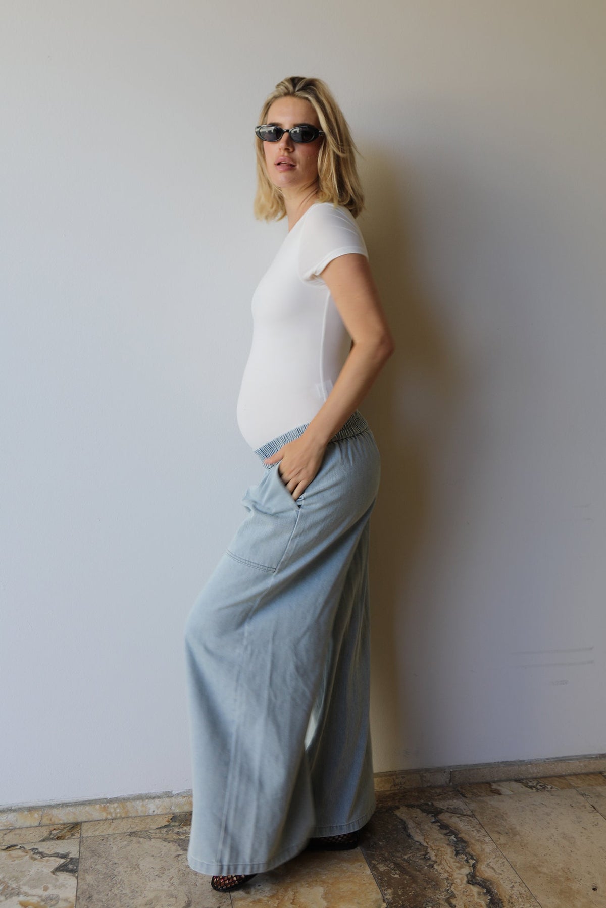 A pregnant woman wearing a white short-sleeve t-shirt and light blue wide-leg denim maxi jeans by Bumpsuits, standing against a plain off-white wall.