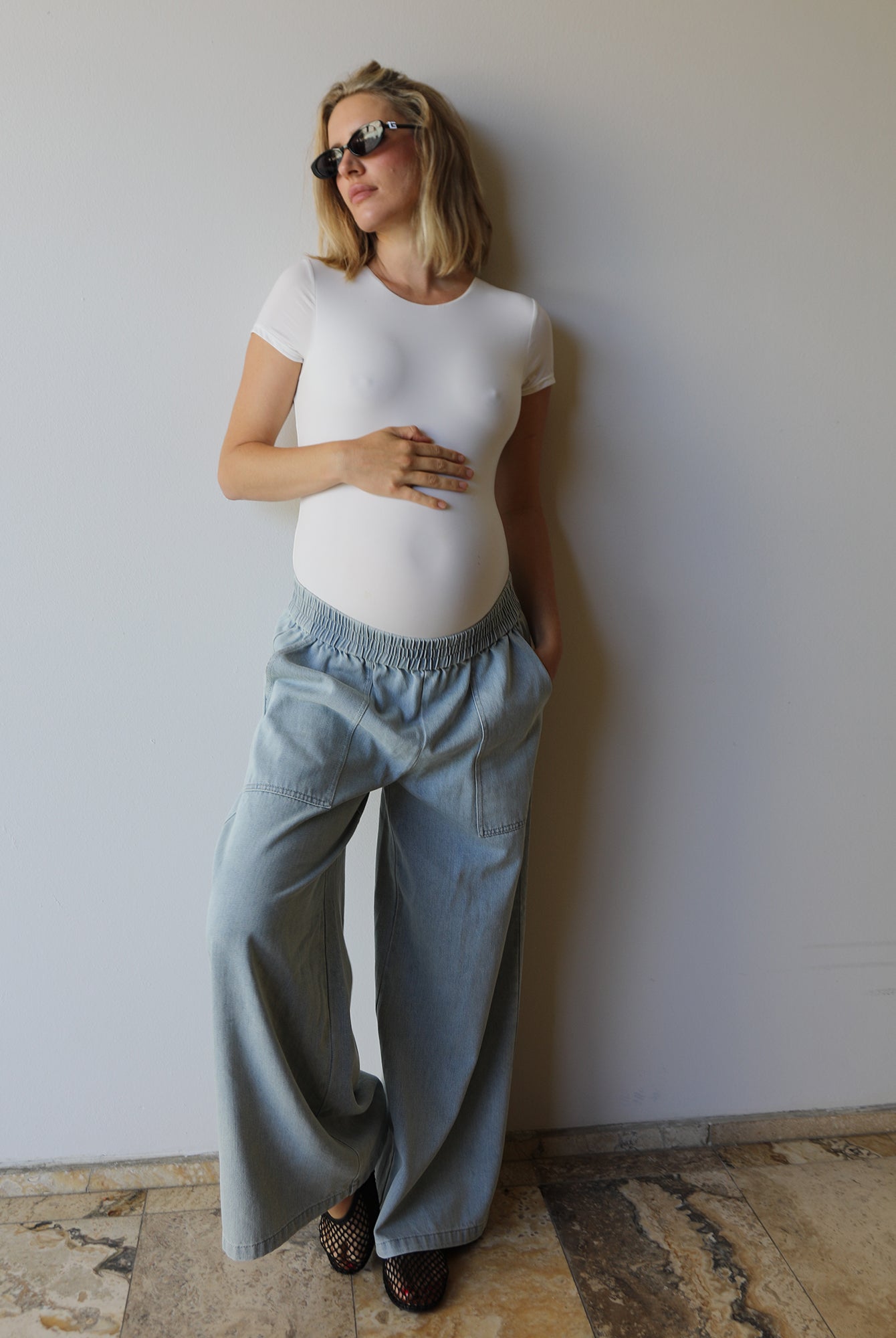 A pregnant woman wearing Bumpsuits white short-sleeve Everyday maternity top and light blue wide-leg jeans, standing against a plain white wall on a stone-tiled floor.