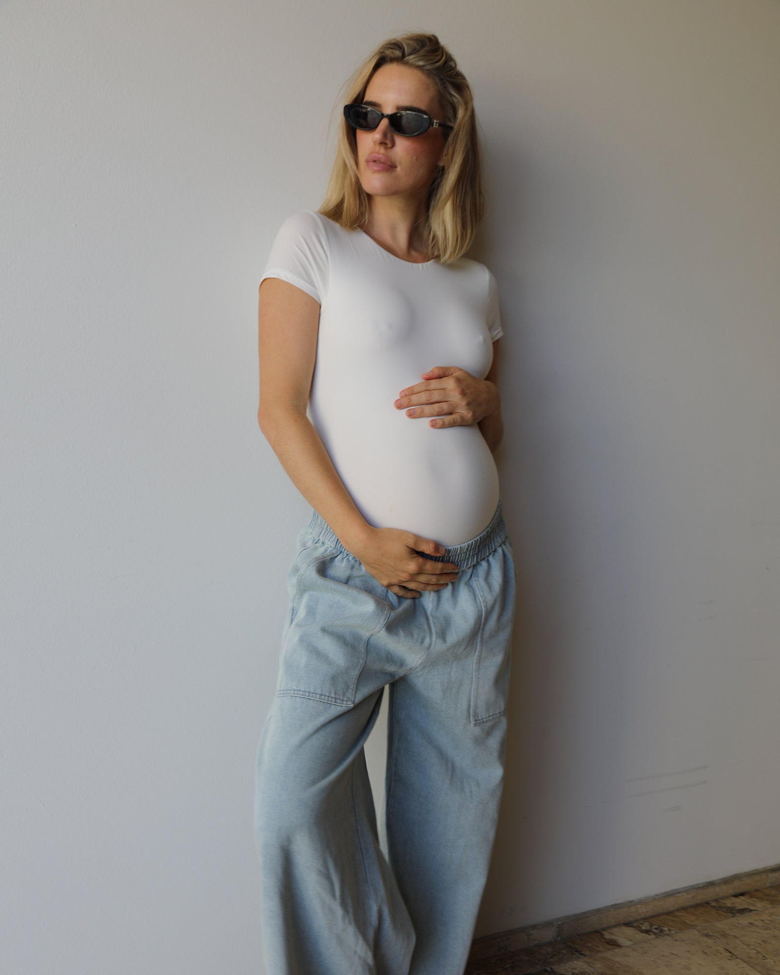 A pregnant woman wearing a fitted white short-sleeve t-shirt and light blue wide-leg jeans by Bumpsuit standing against a plain light gray wall.