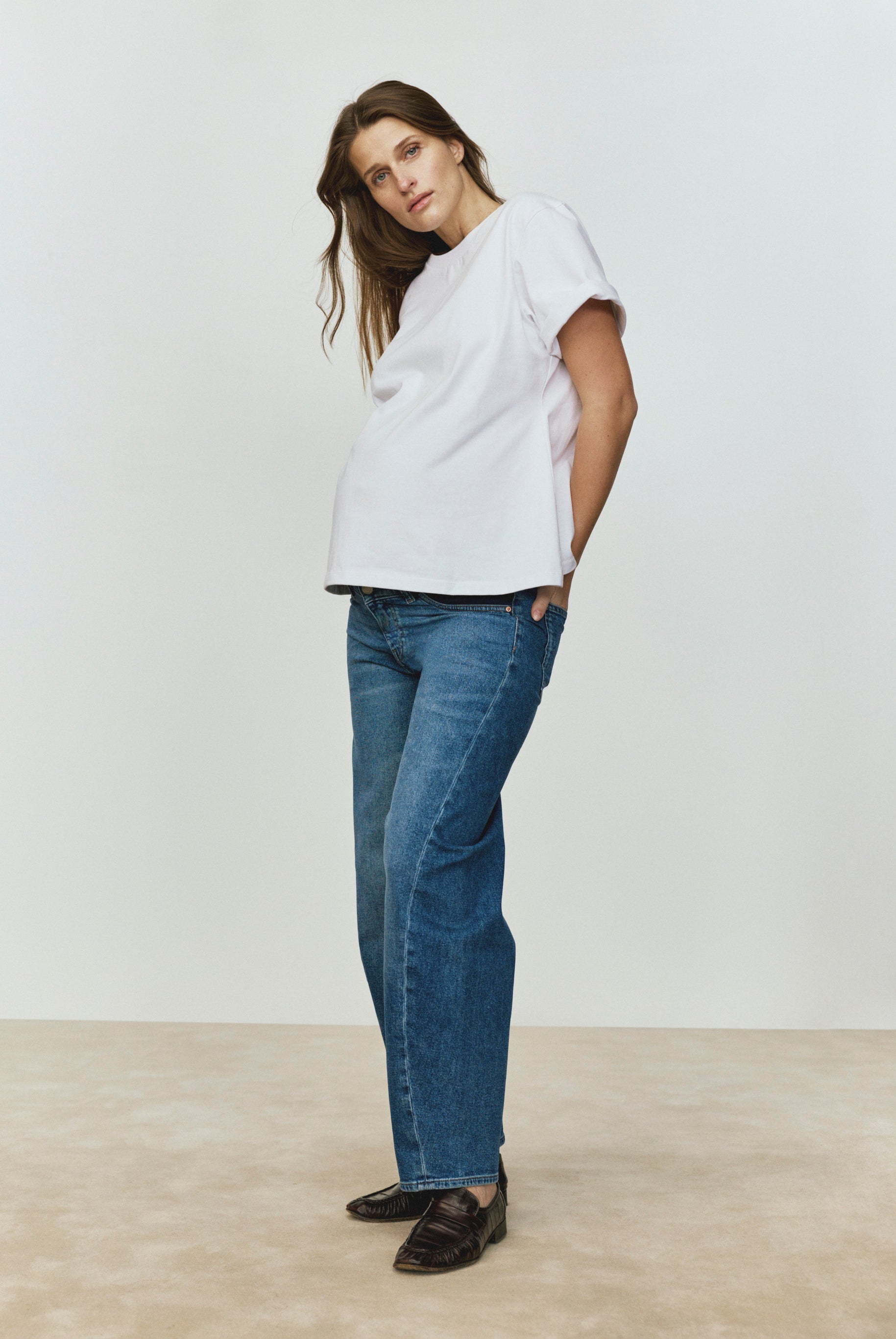 Pregnant woman wearing a white top and blue jeans standing against a plain background
