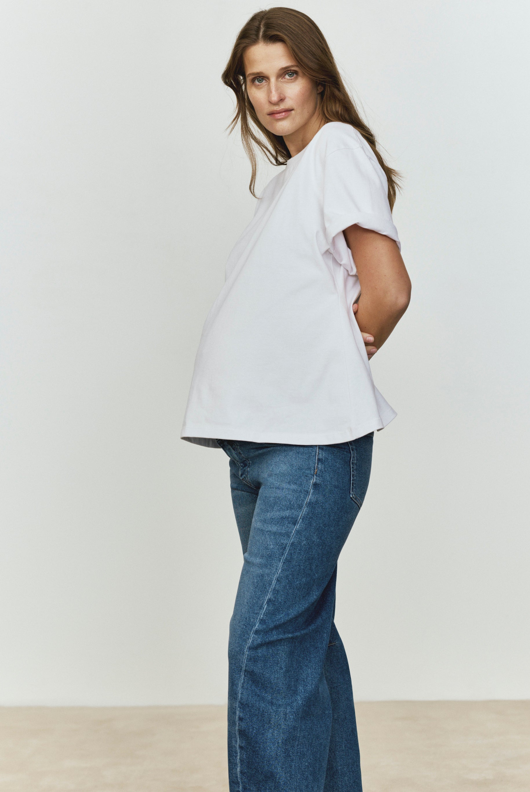 Pregnant woman wearing a white t-shirt and blue jeans against a plain background